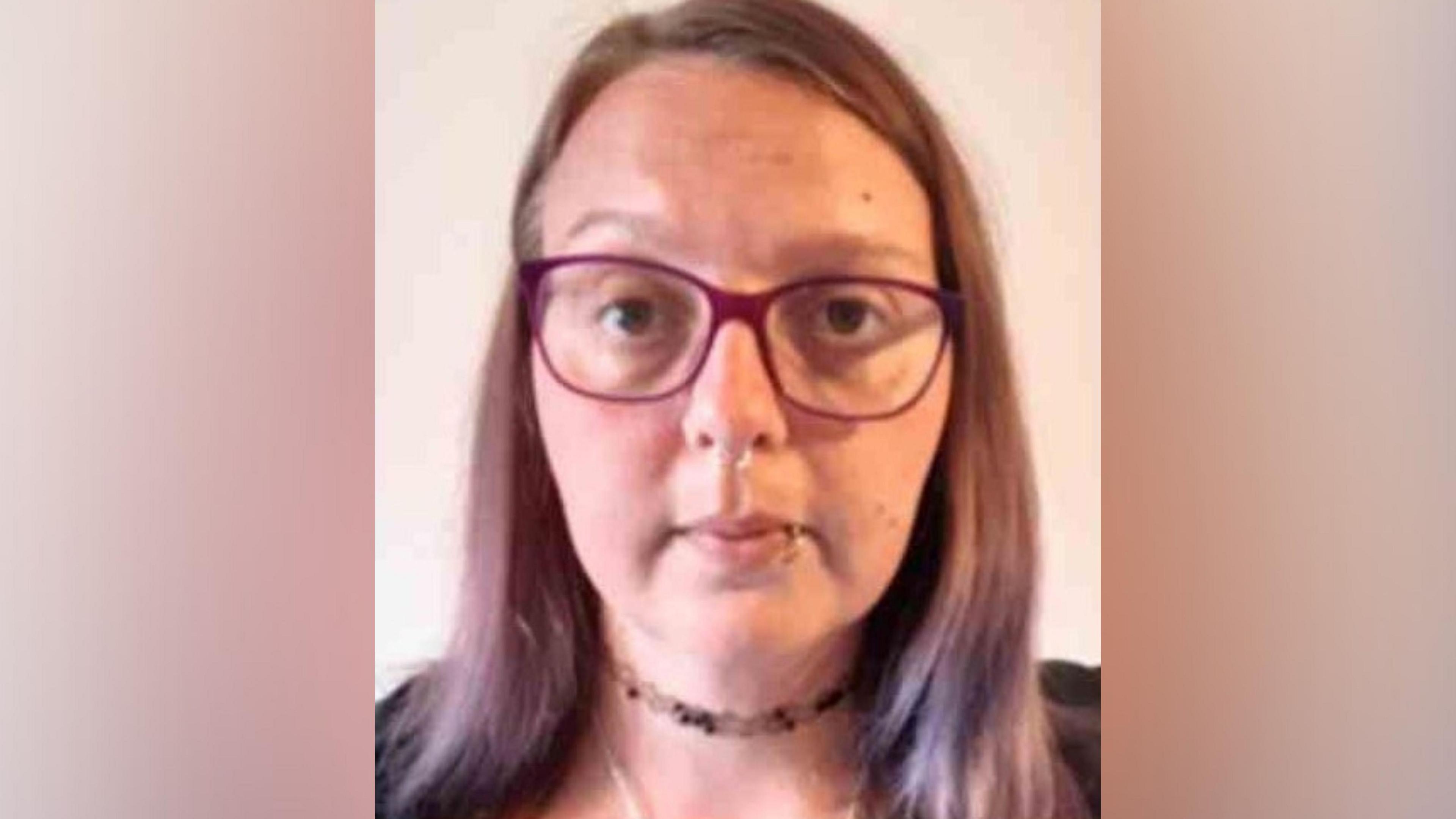 Custody photograph of Rebecca Welch who is wearing glasses with a purple frame, has nose and lip piercings, and a choker round her neck. She has light purple hair which is just over shoulder length 