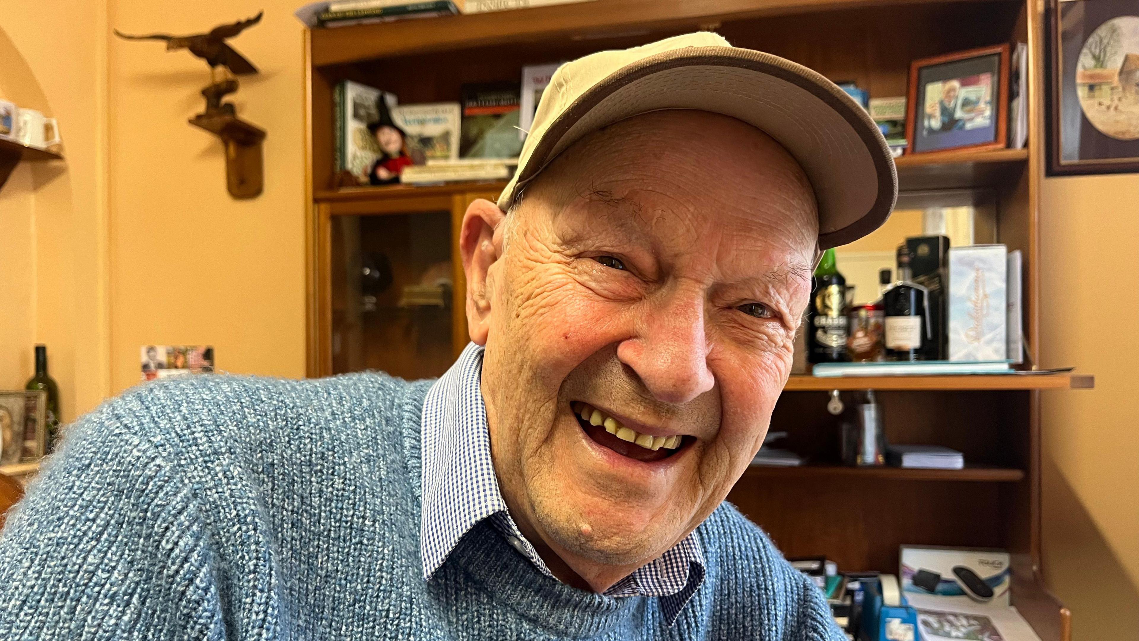 Malcolm Metcalf laughs at the camera. He is in a room with a cabinet behind him holding photographs and office paraphernalia. There are pictures on the wall and a statue of a bird on the wall.