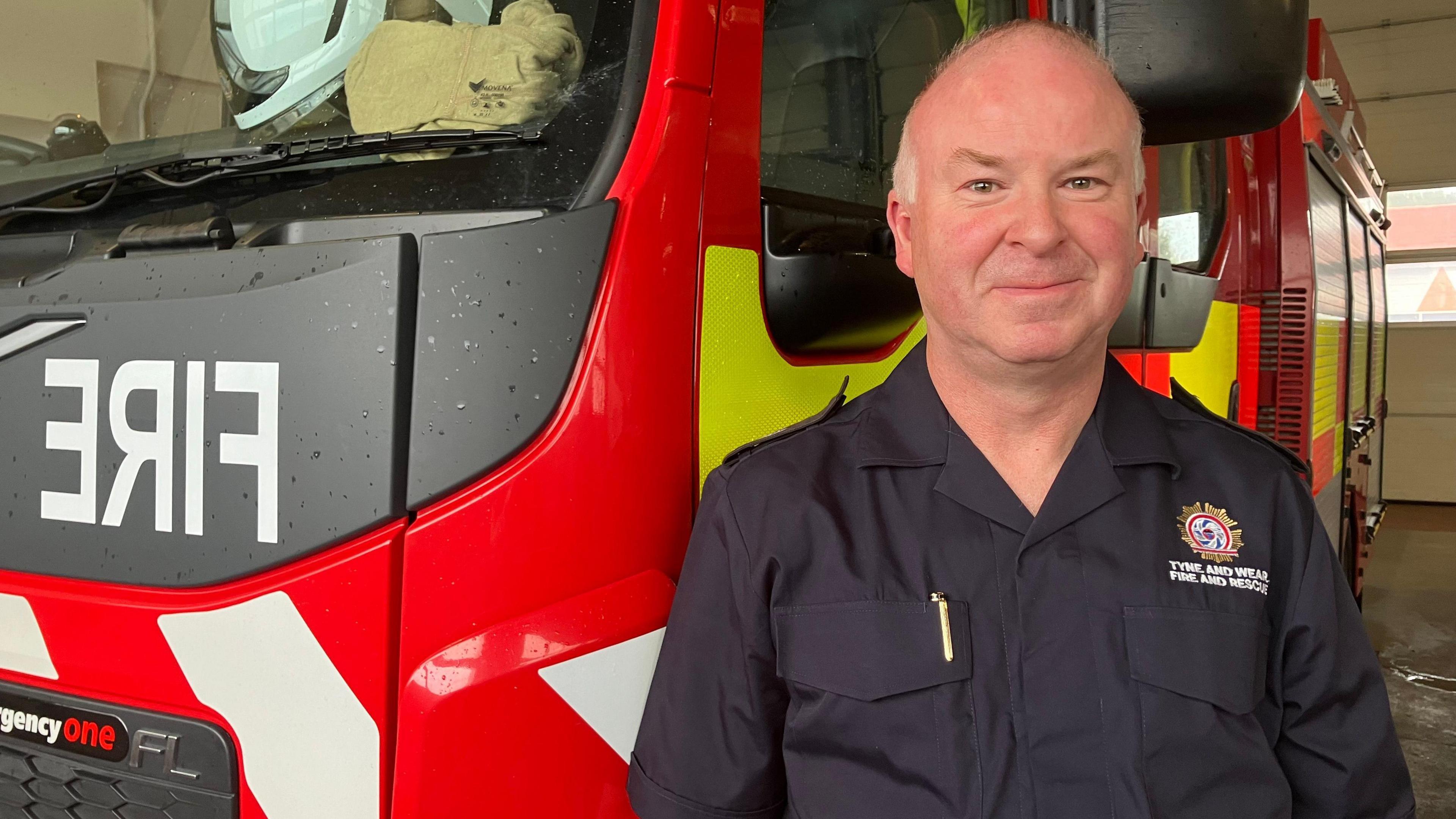 Phil Clark is stood next to a fire engine. He is looking directly at the camera is wearing a Tyne and Wear Fire and Rescue Service uniform. 