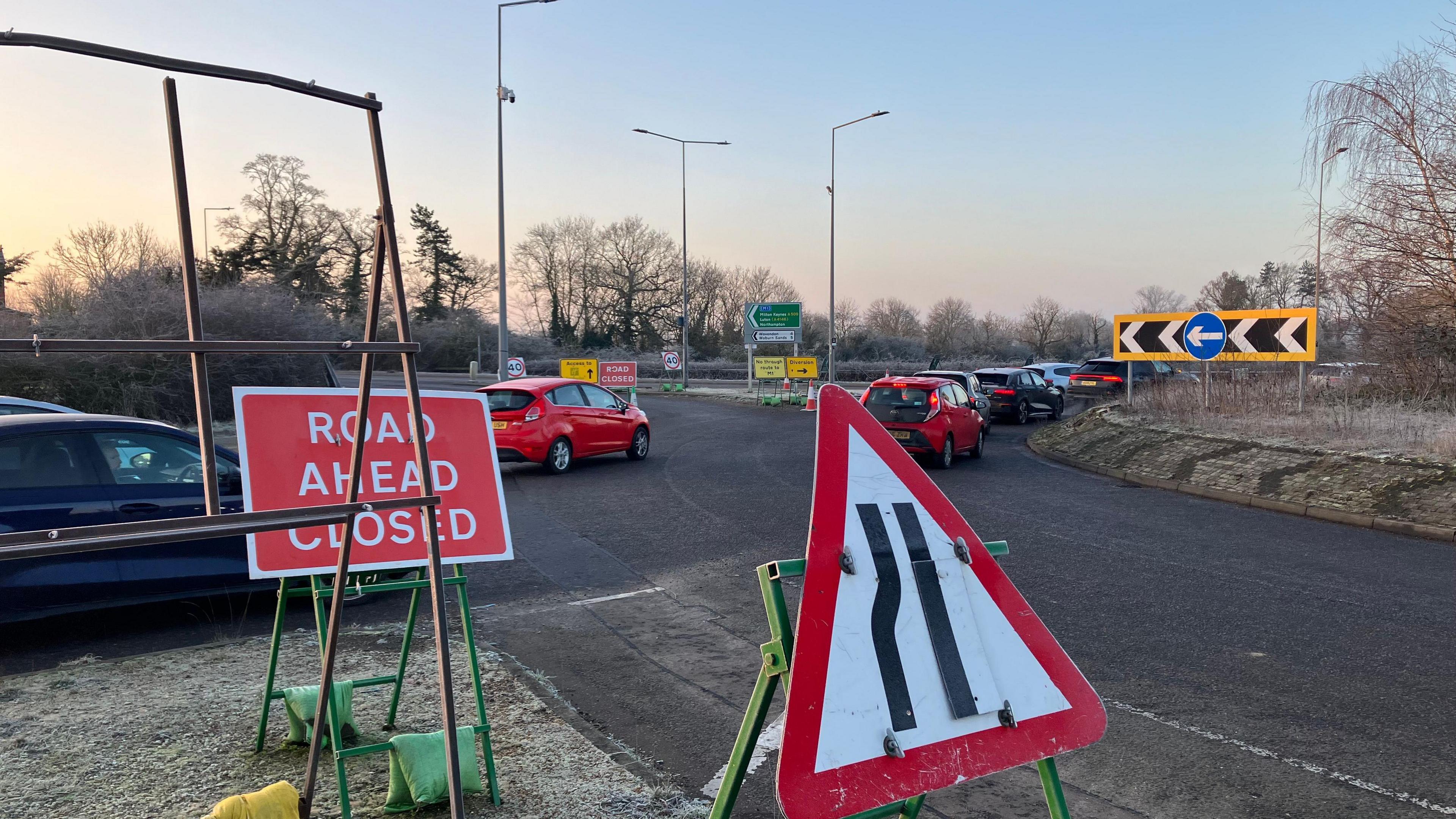 A picture of a roundabout with cars on it, stuck in a queue. There are also road signs to indicate that a nearby road is closed.