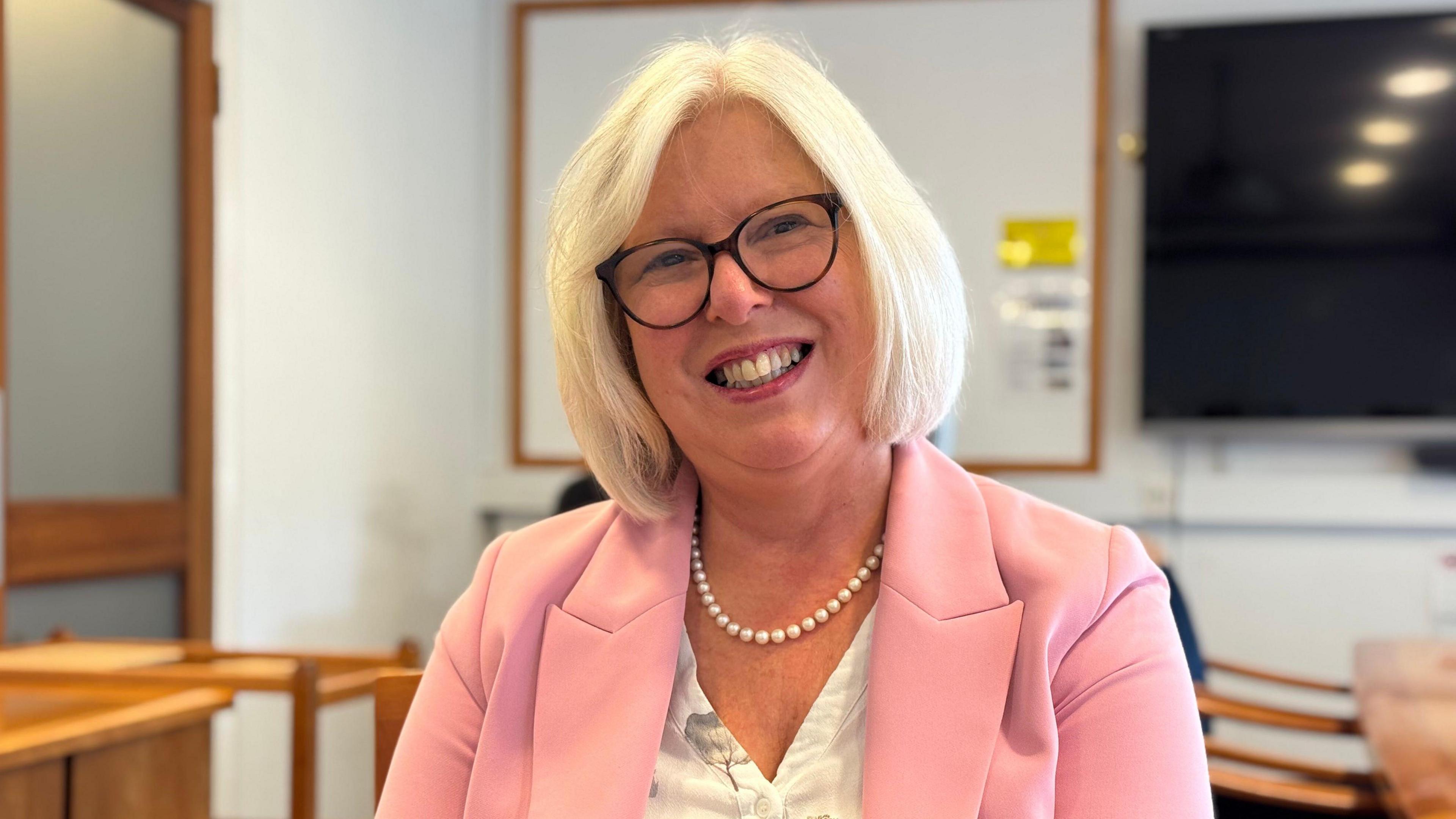 Dr Nicola Brink, who has blonde hair, large black glasses and wearing a pink jacket, is seen smiling at the camera. Behind her is a TV screen and part of a desk is visible.