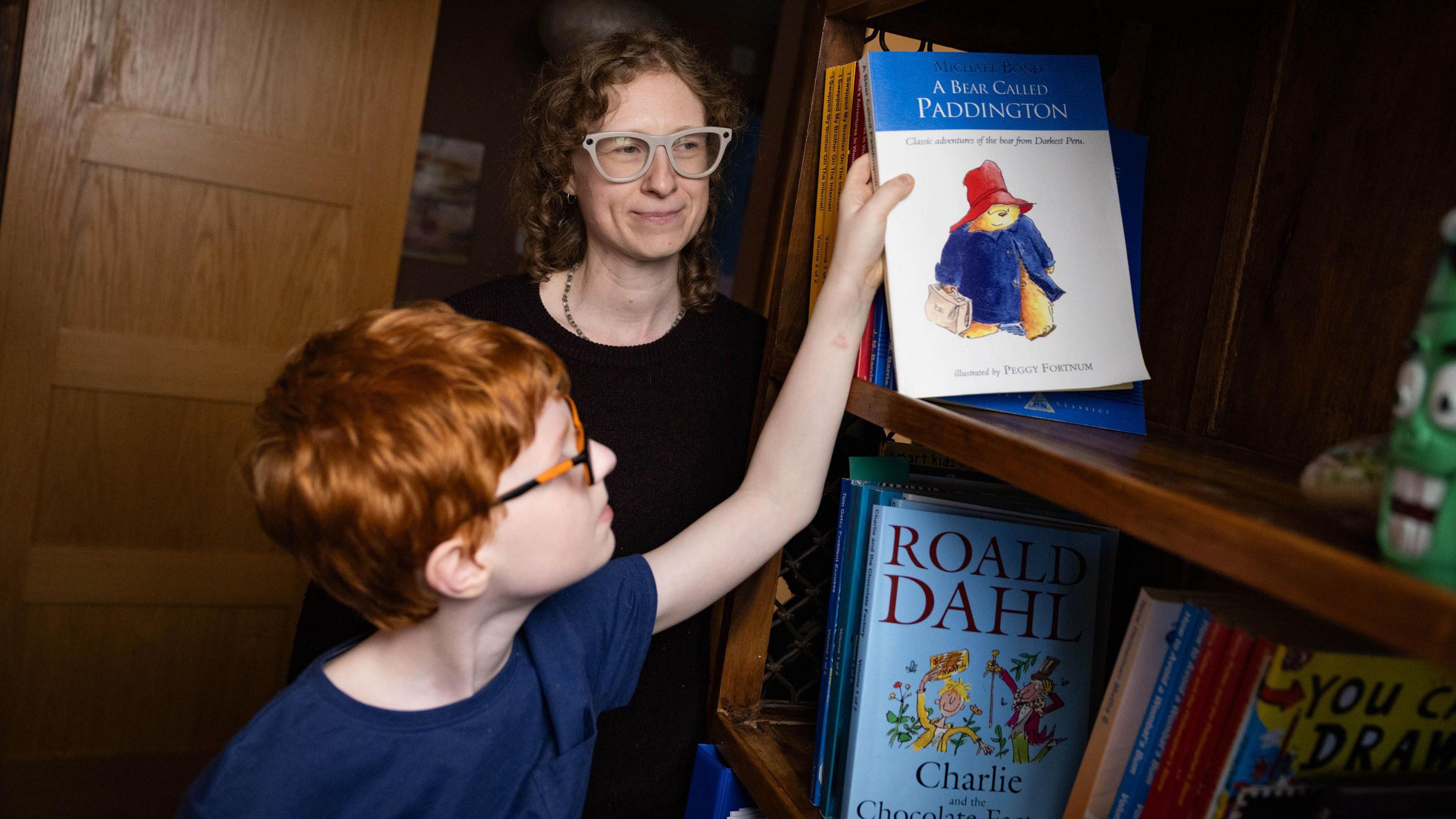 A boy with red hair reaches up to a book shelf to grab a book. His mum is smiling in the background