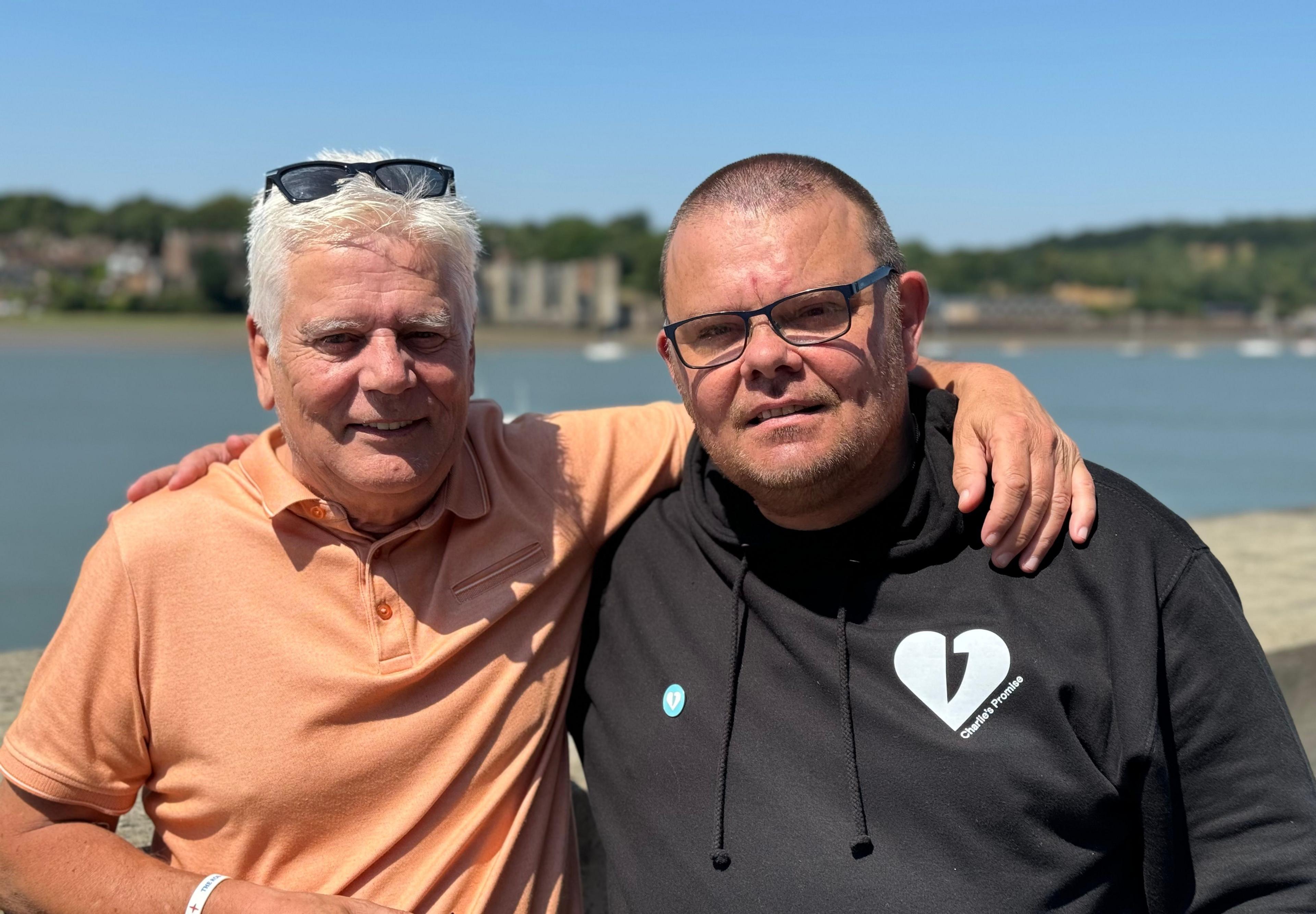 Colin Knox, wearing a peach coloured top has his arms over the shoulder of Martin Cosser, who is wearing a black top with a heart symbol