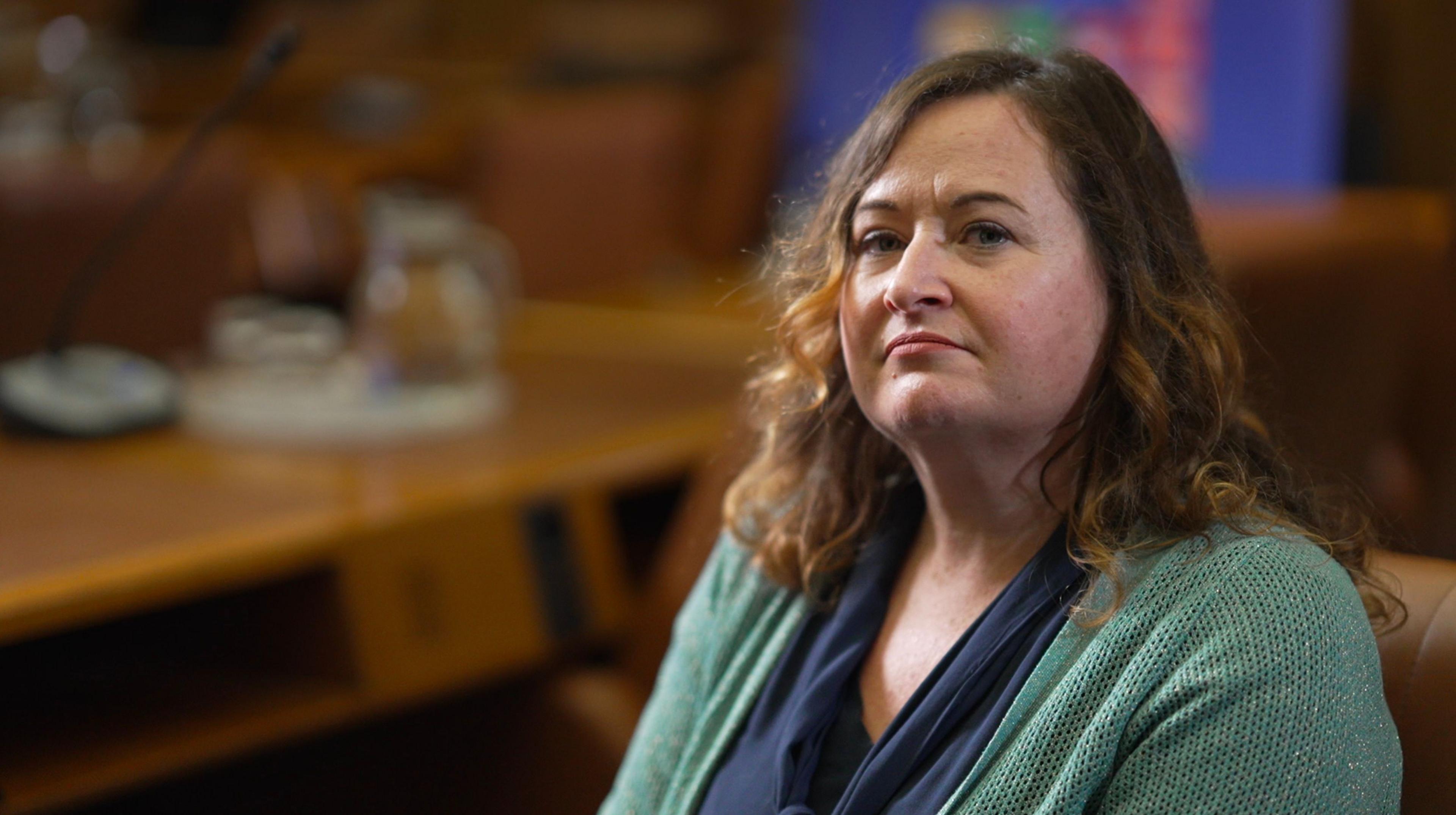 Claire Healy of the UN’s Office on Drugs and Crime (UNODC), sitting in an office looking at the camera, with a desk behind her. She is wearing a green cardigan and a blue top underneath. Her brown hair with blonde highlights is untied.