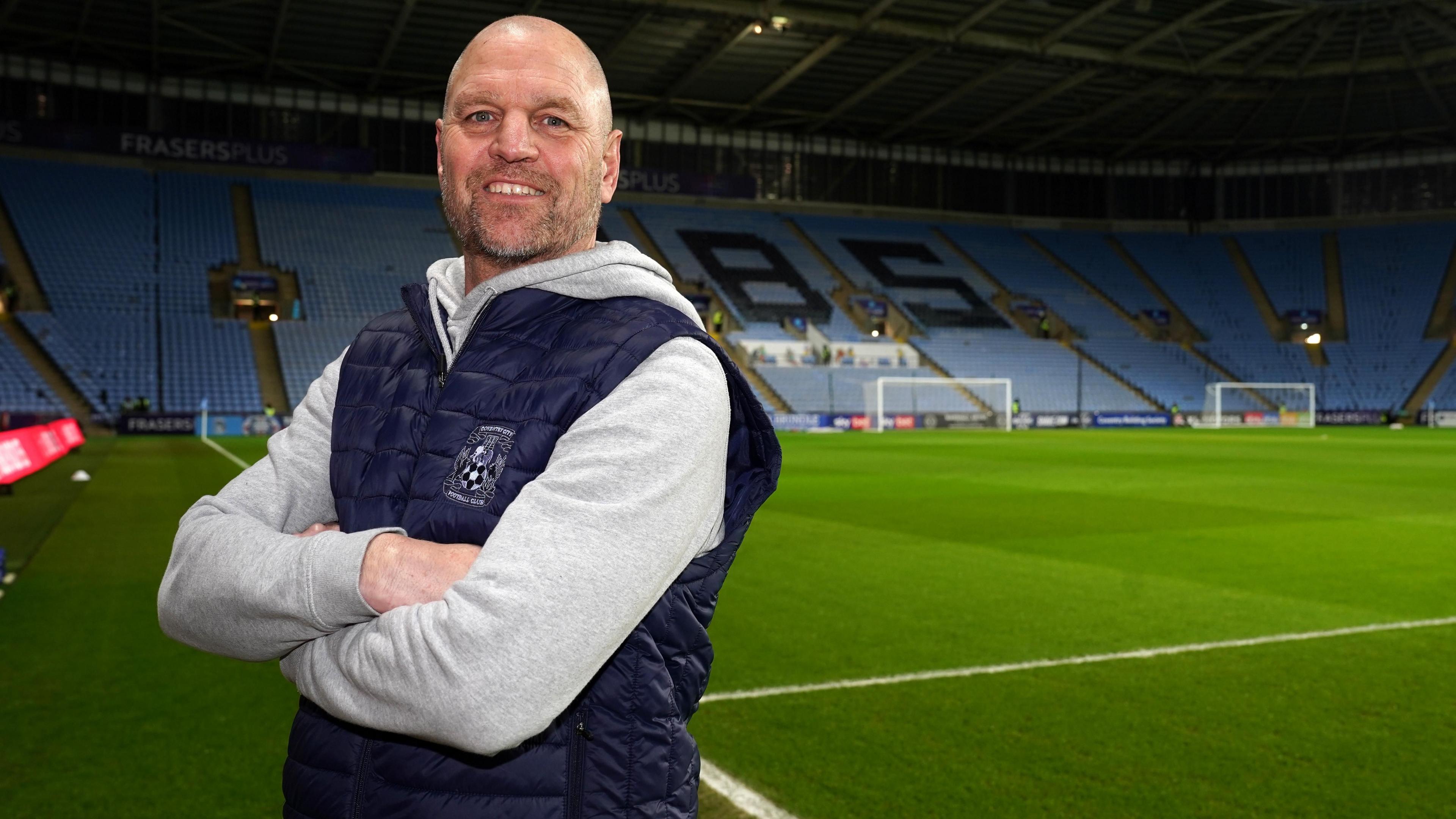 A man with a shaved head and stubble, wearing a grey hoodie and a sleeveless jacket with the Coventry City FC logo on it, stands on a football pitch smiling at the camera.