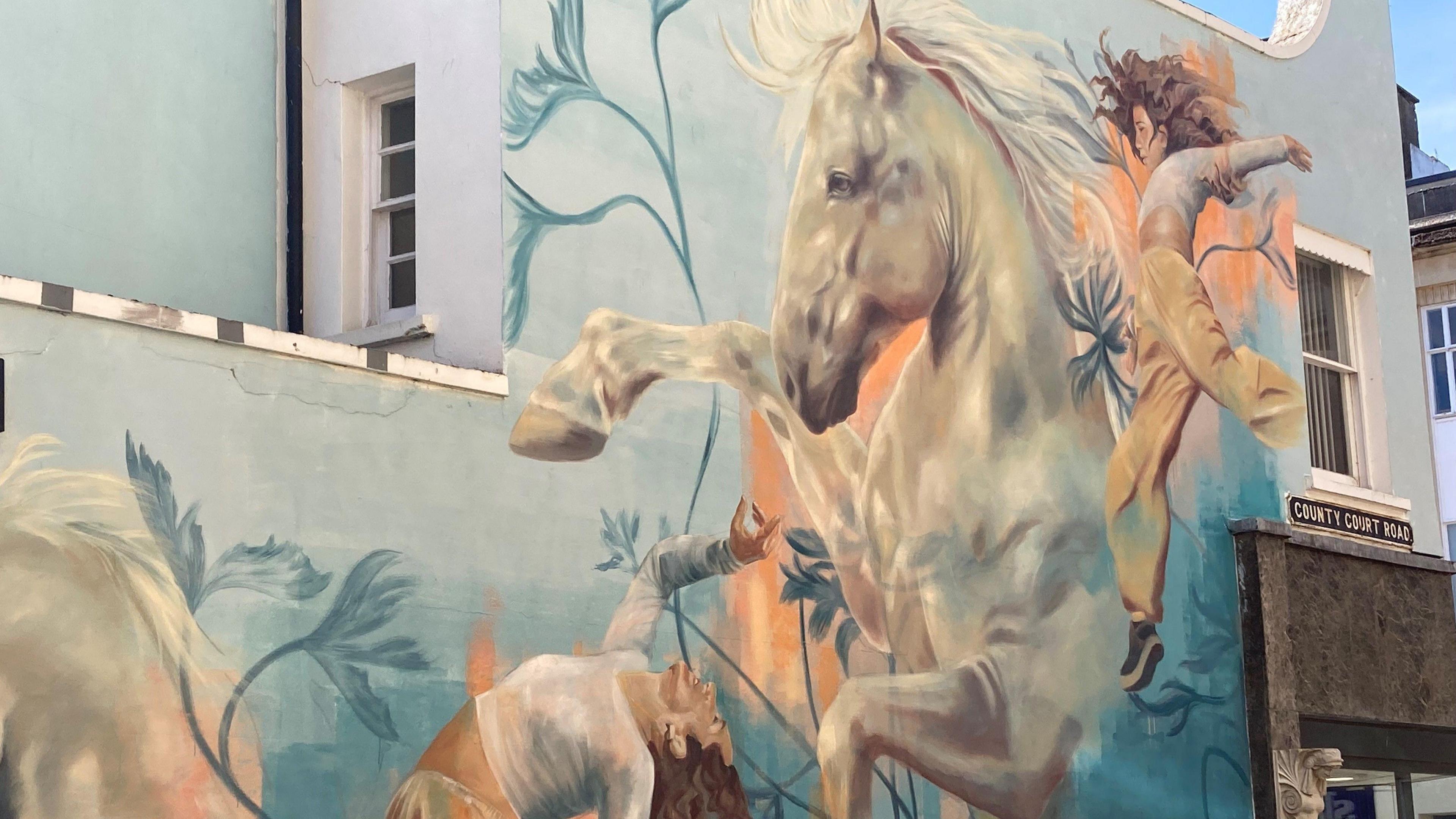 A mural showing a white horse with women dancing around it on the side of a building