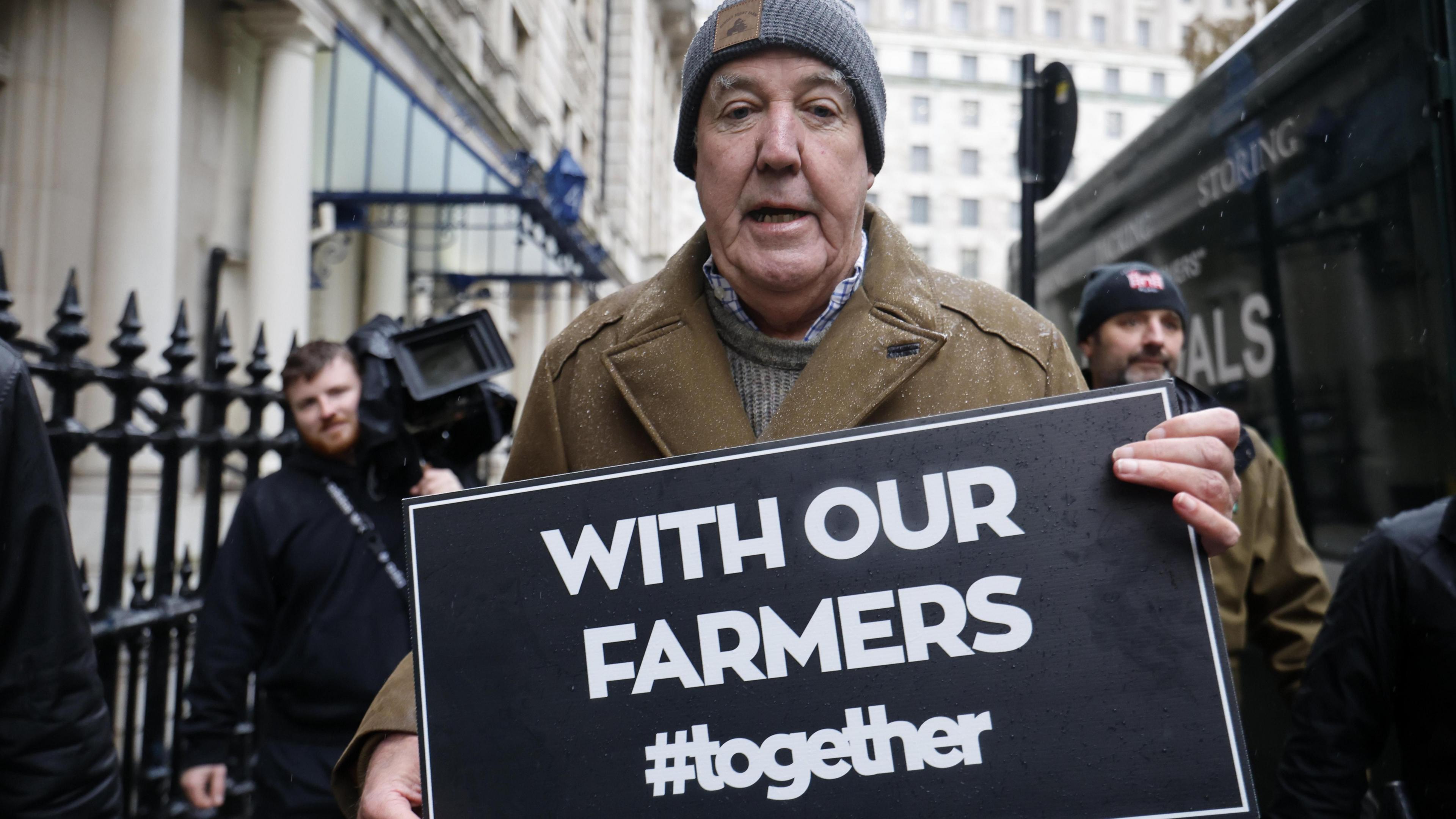 Jeremy Clarkson wearing a beige coat and grey hat, carrying a placard saying With Out Farmers #together