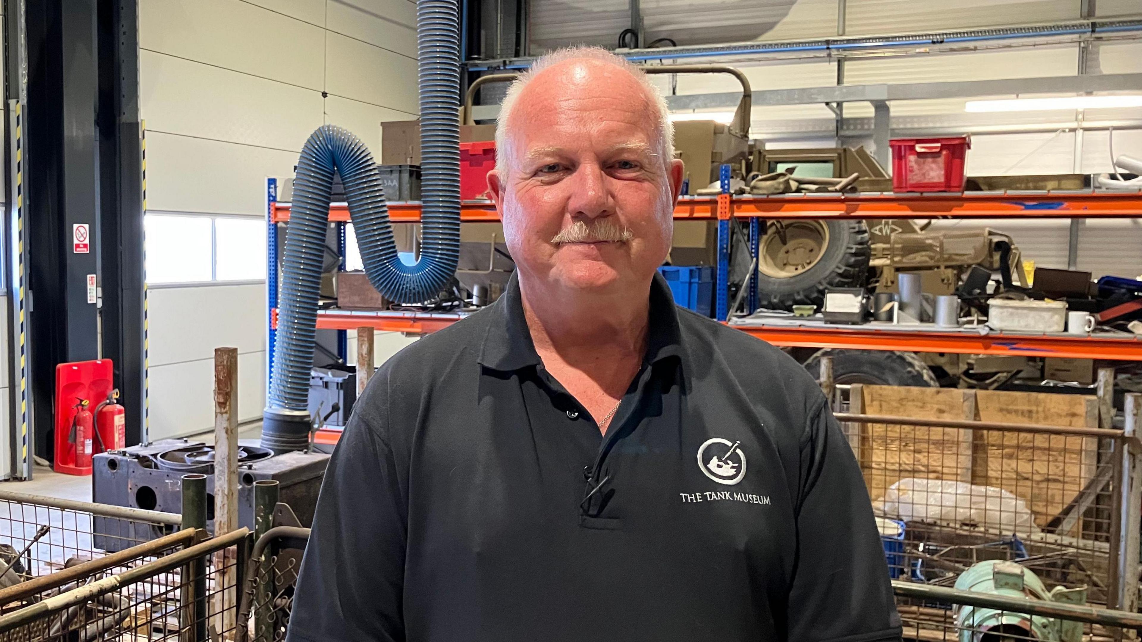 Duncan Masters, Workshops Manager, The Tank Museum stands in the workshop. He is wearing a black Tank Museum t-shirt. The background is full of all sorts of bits and pieces that have come off various tanks.