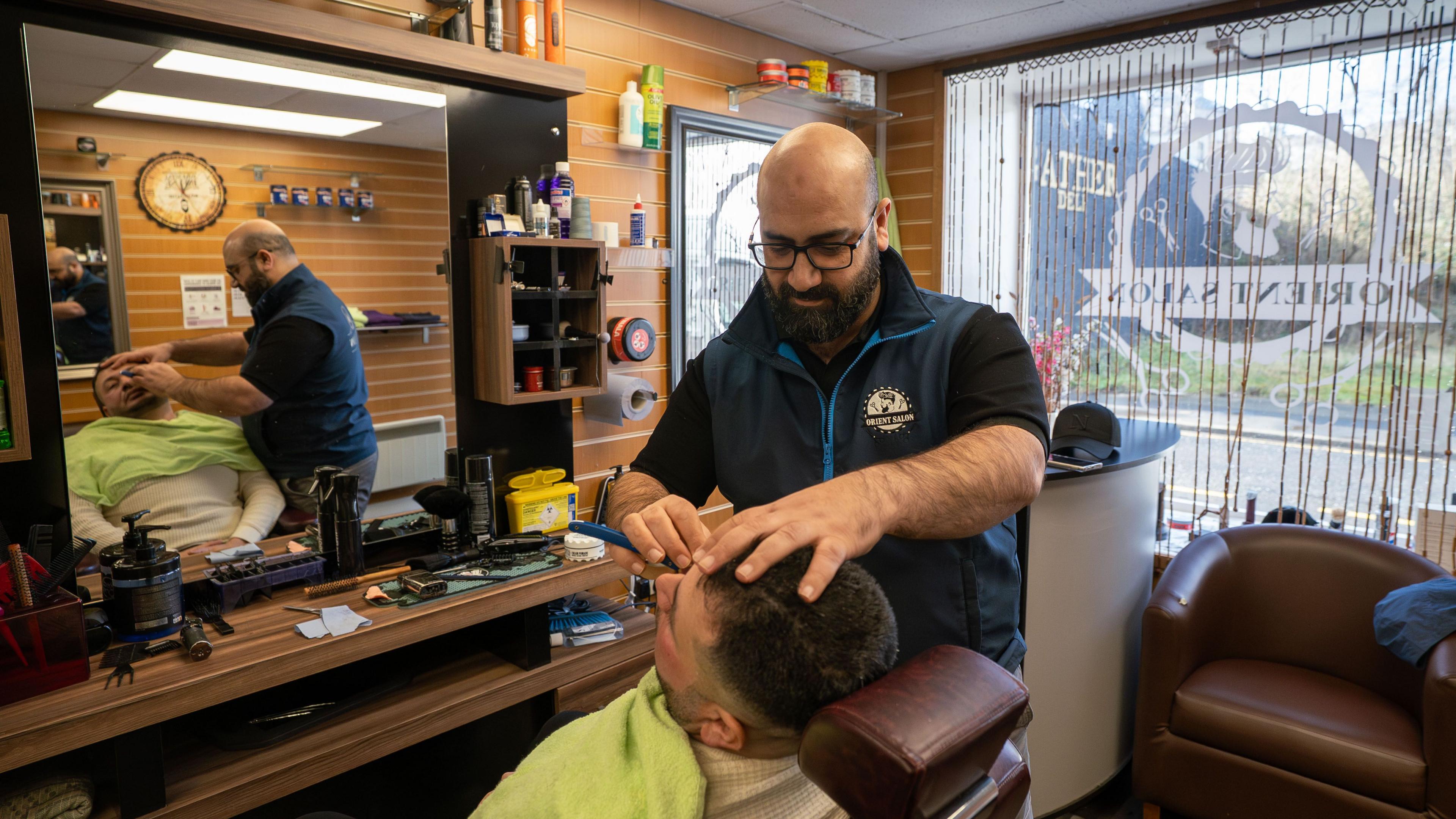 Mounzer Darsani, a bald man with dark-rimmed glasses and a black beard, shaves a man's face in his barbershop. He is wearing a blue body warmer over a black short-sleeved shirt. The customer is sitting in a barber's chair with a towel around his shoulders. They are both reflected in the mirror beside them. 