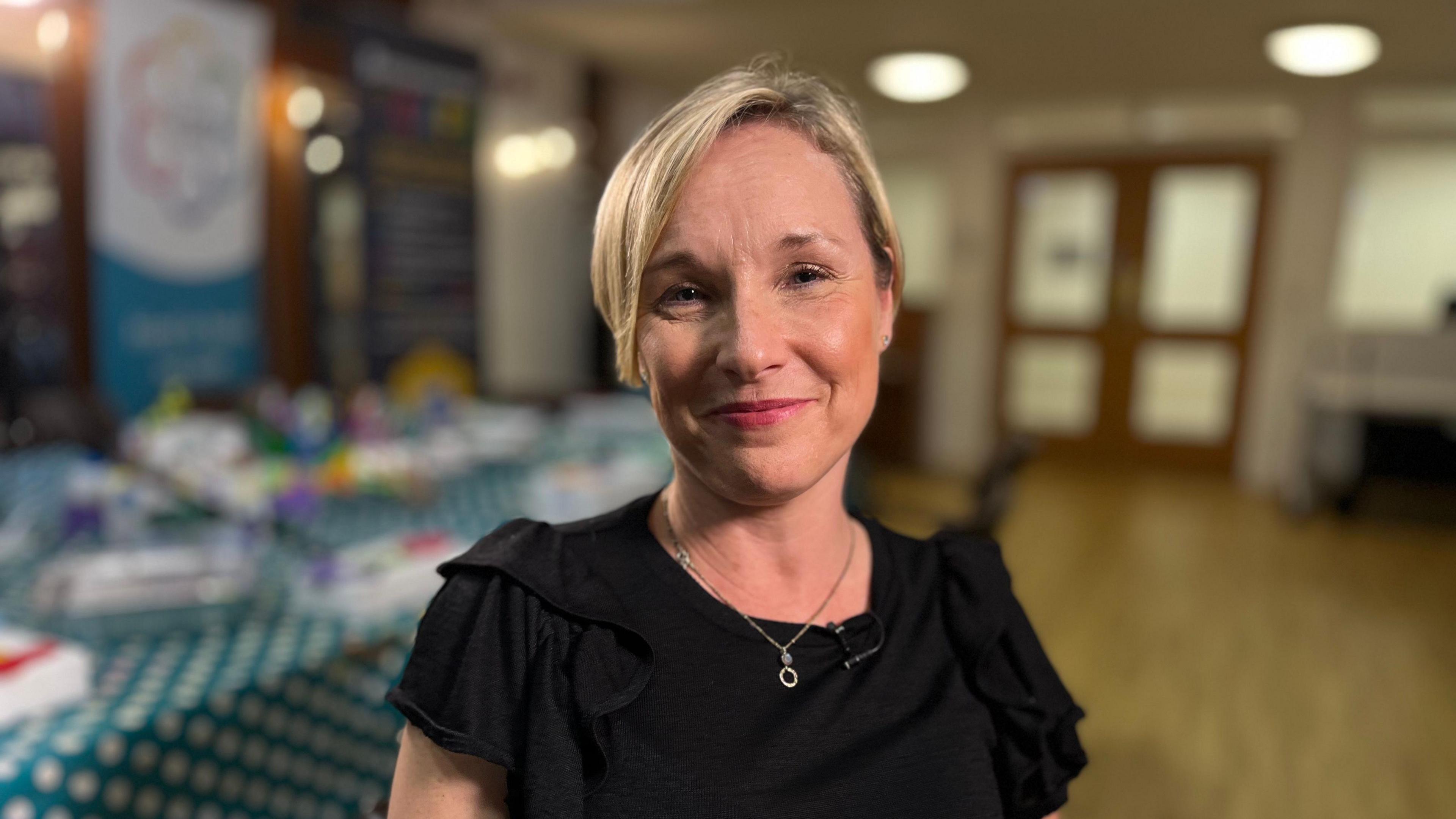 A smiling Amanda Orange looks directly at the camera. She is standing in a hall, and is wearing a black blouse and silver necklace. 