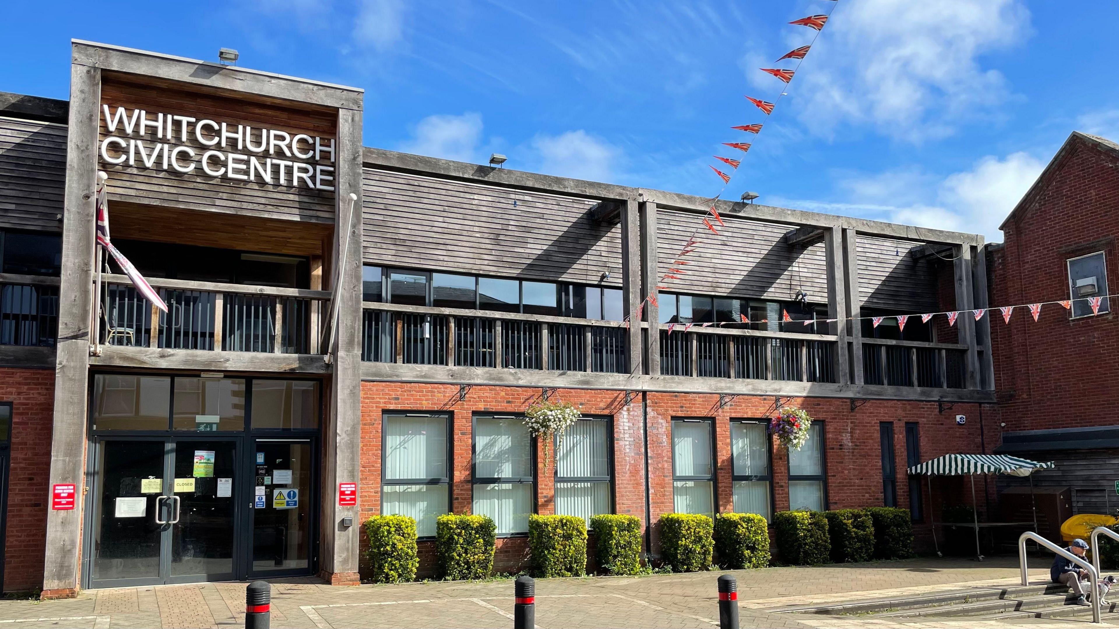 A two-storey building with a dark oak balcony on the first floor. The ground floor is red-bricked, with a glass double door to the left and six tall windows to the right. The sky is blue and cropped bushes run along the front of the building.
