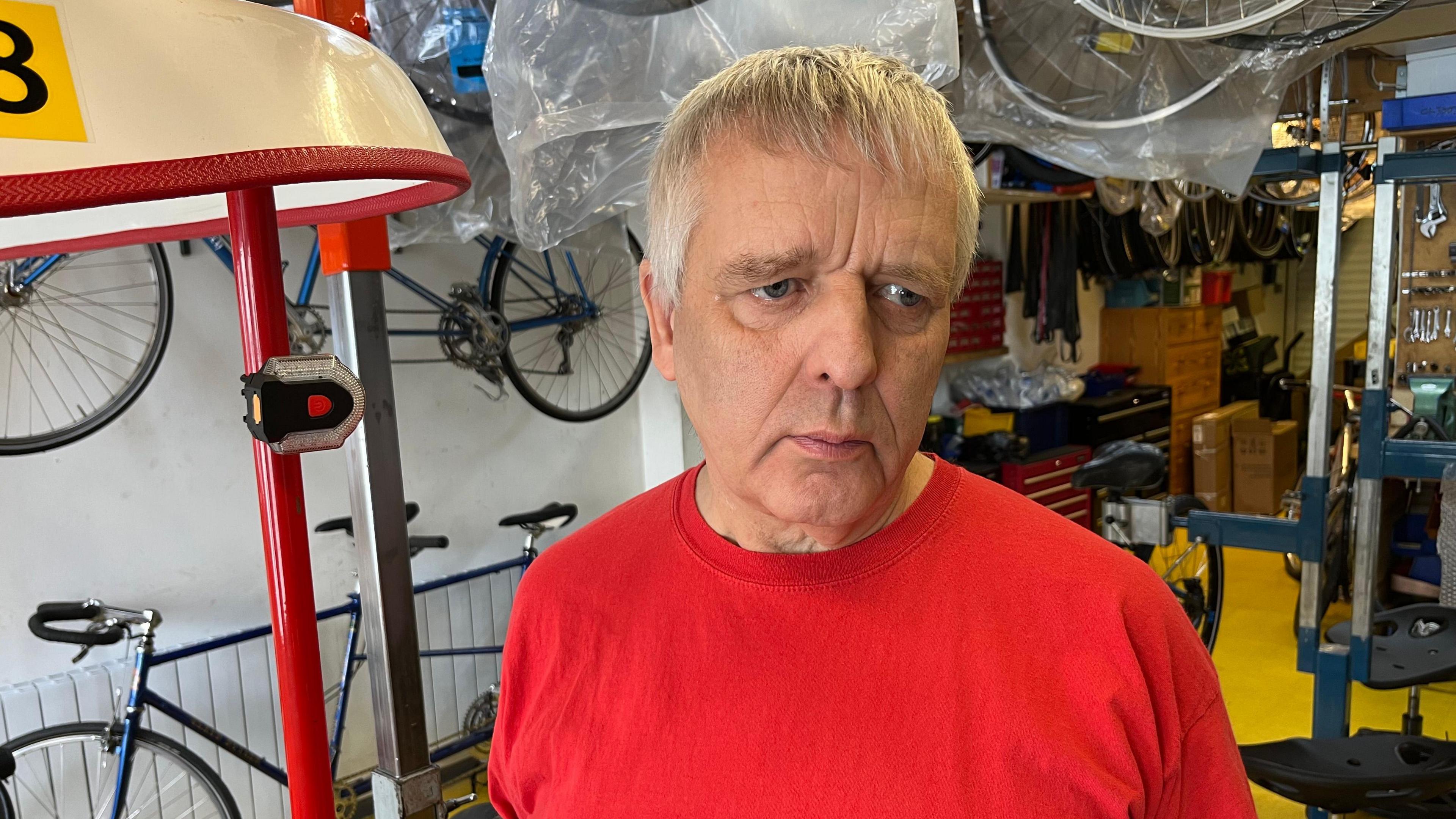 Graham Hicks, 64, has short grey hair with a fringe near the top of his head, he is wearing a red T-shirt and behind him are various tools and machines for mending bikes and some bike frames too.