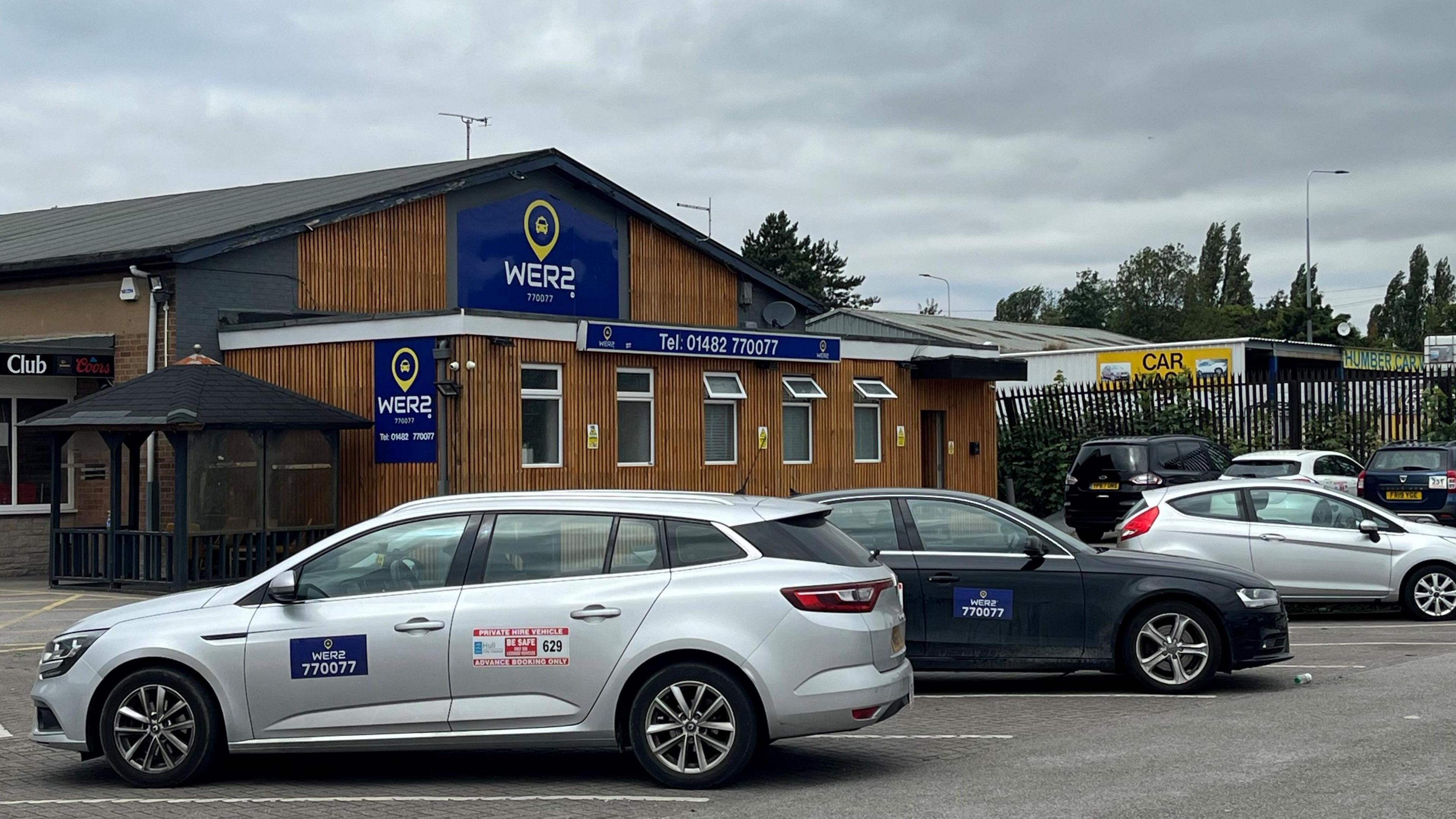 The Wer2 taxi depot, which has blue signs and wooden cladding. Silver and back private-hire taxis are parked outside