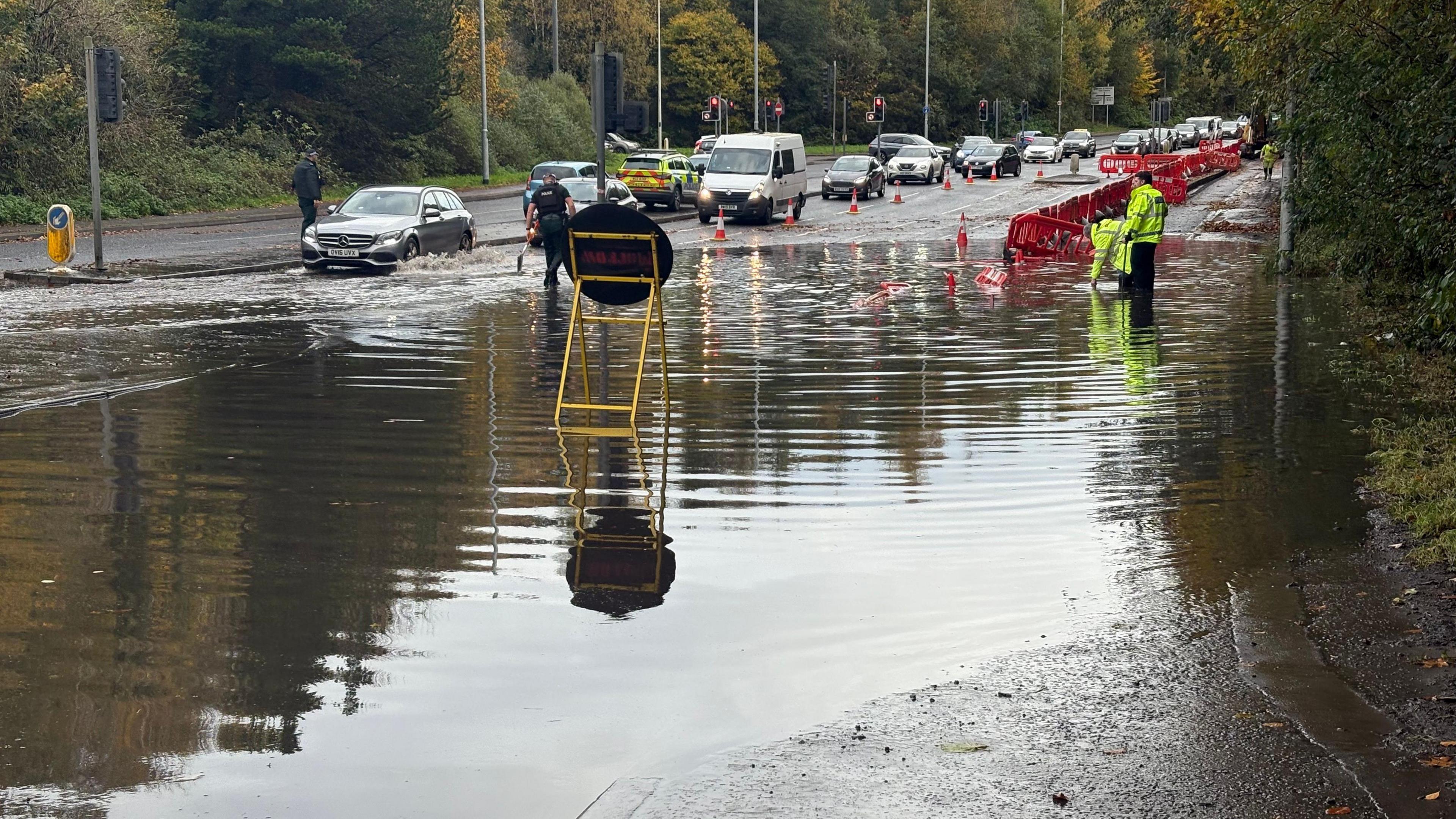 People in high-viz standing in a large puddle with red barriers. PSNI officers are directing a line of traffic.