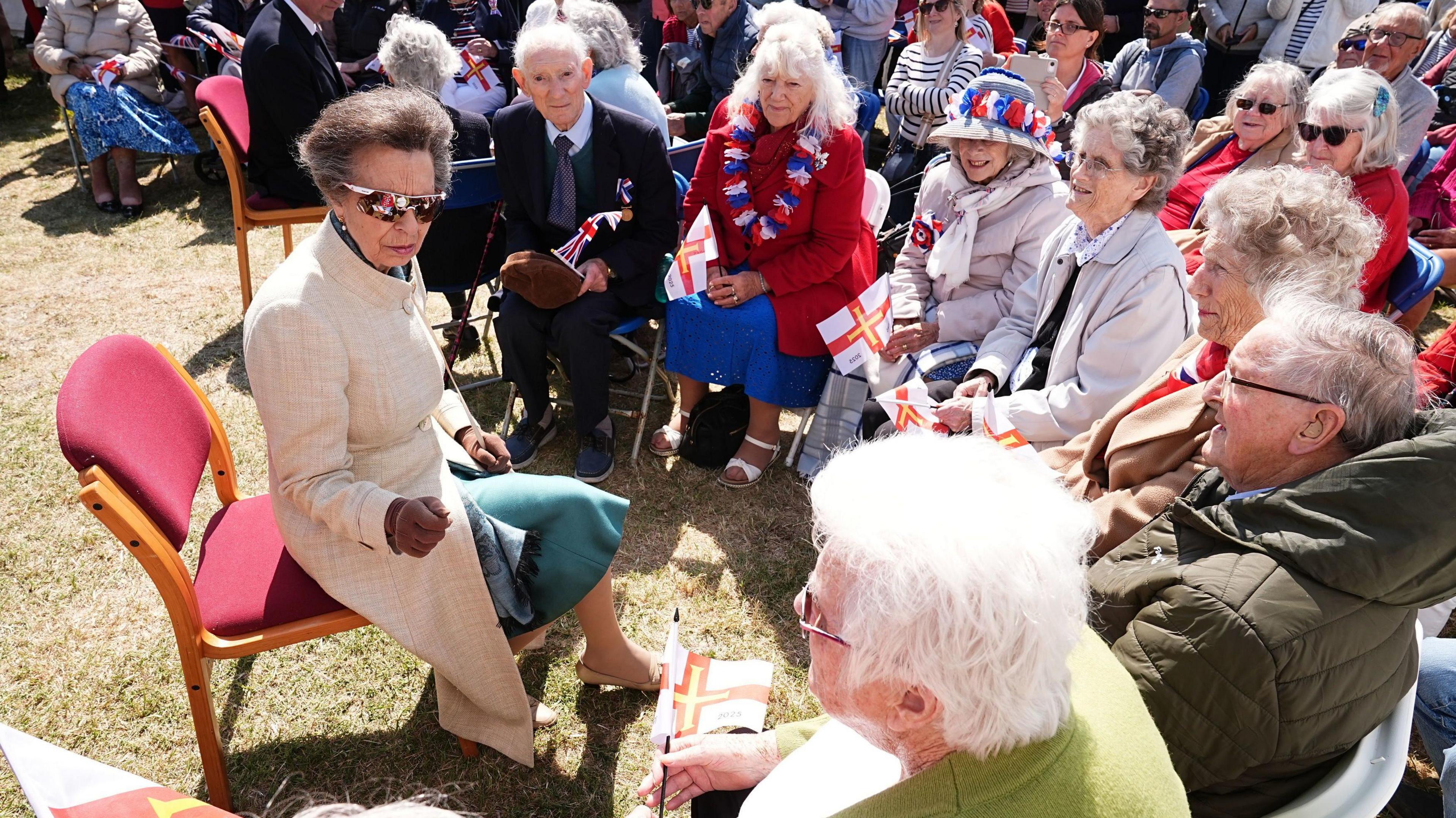 A picture of Anne sat on a red chair surrounded by a circle of people. They are all smiling and speaking with Anne who is wearing gloves and glasses.