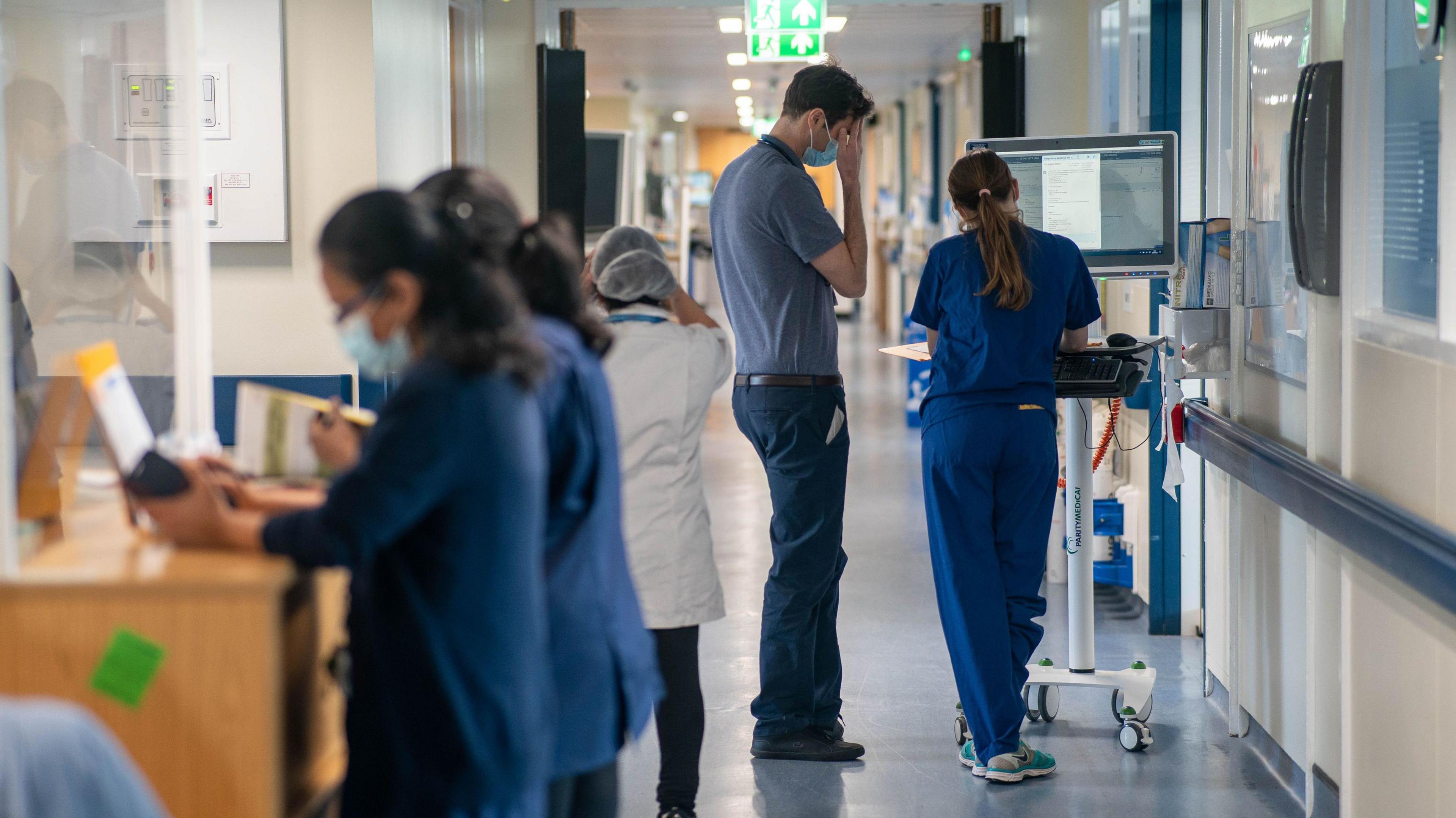 Two medical staff stand in front of a monitor in hospital corridor while more two more staff stand at the side at a reception desk. 