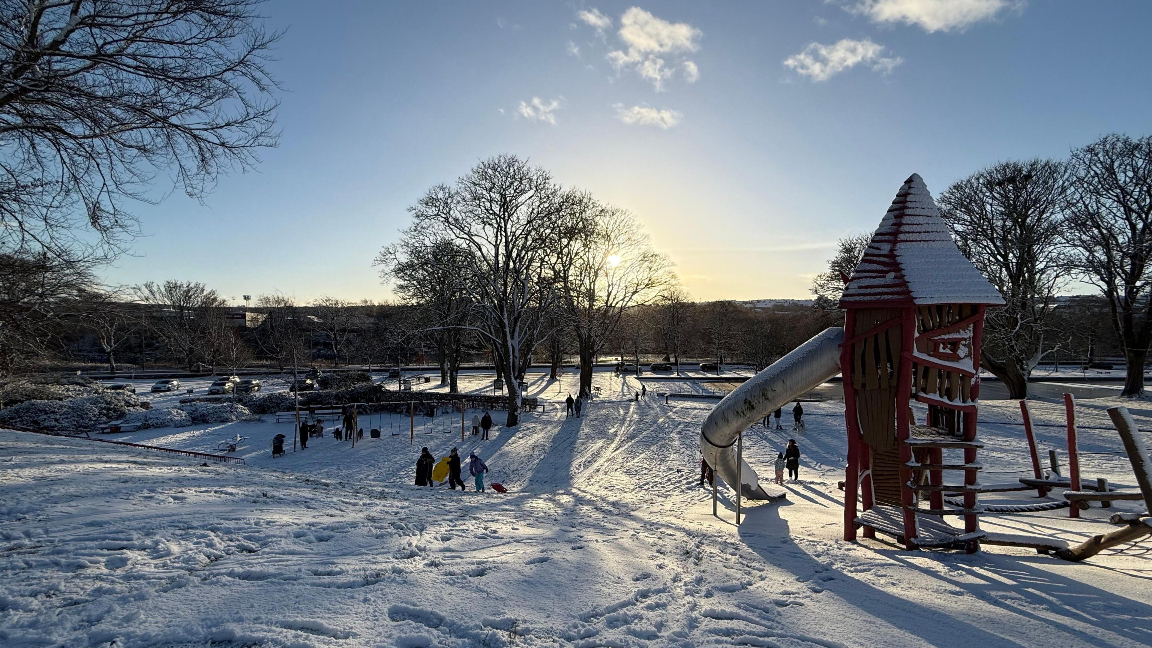 Snow on ground in playground, can see a slide, swings and families playing.