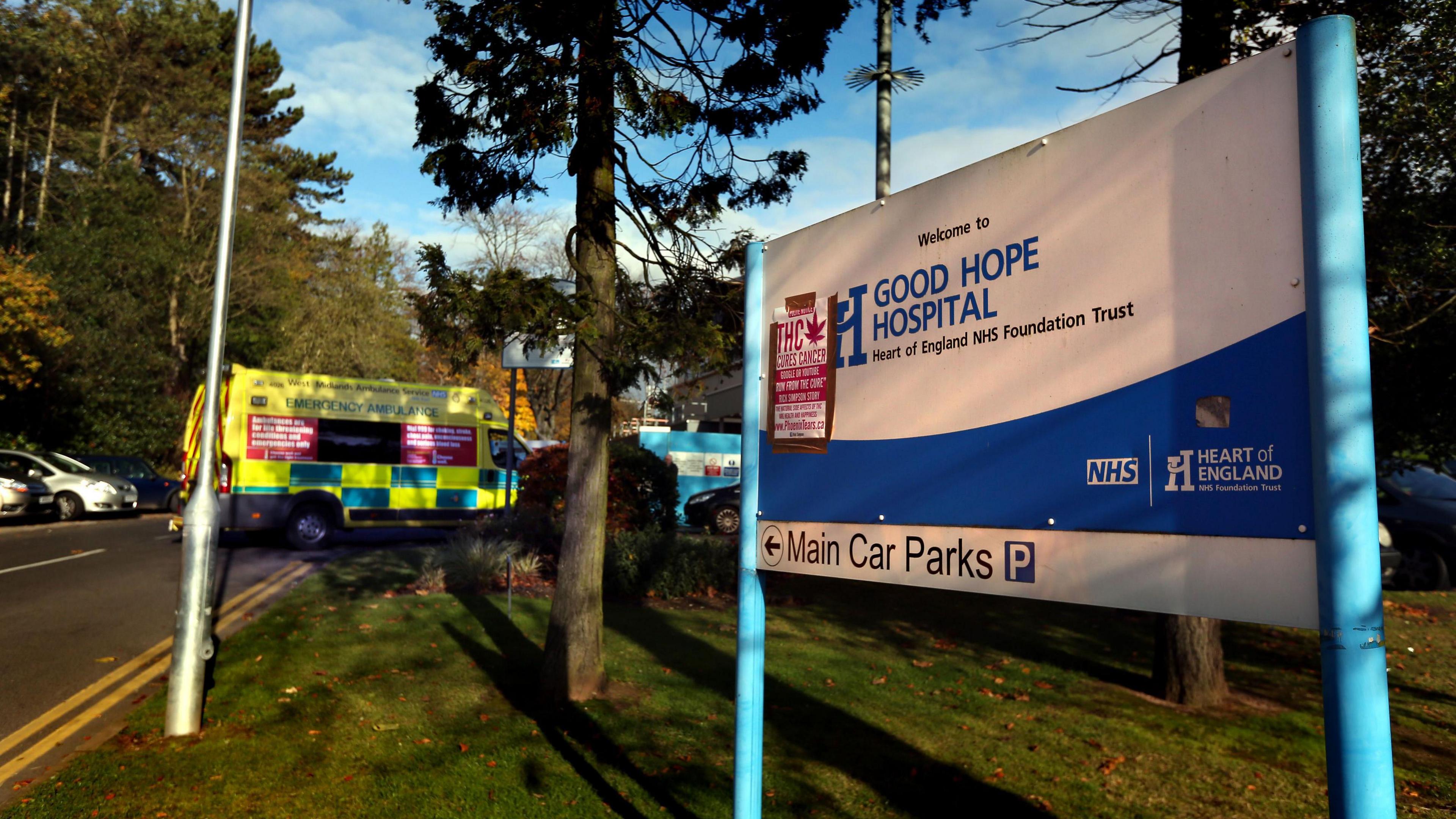 A blue and white metal sign that says Welcome to Good Hope Hospital. In the background a tree is visible in front of an ambulance along a small stretch of road