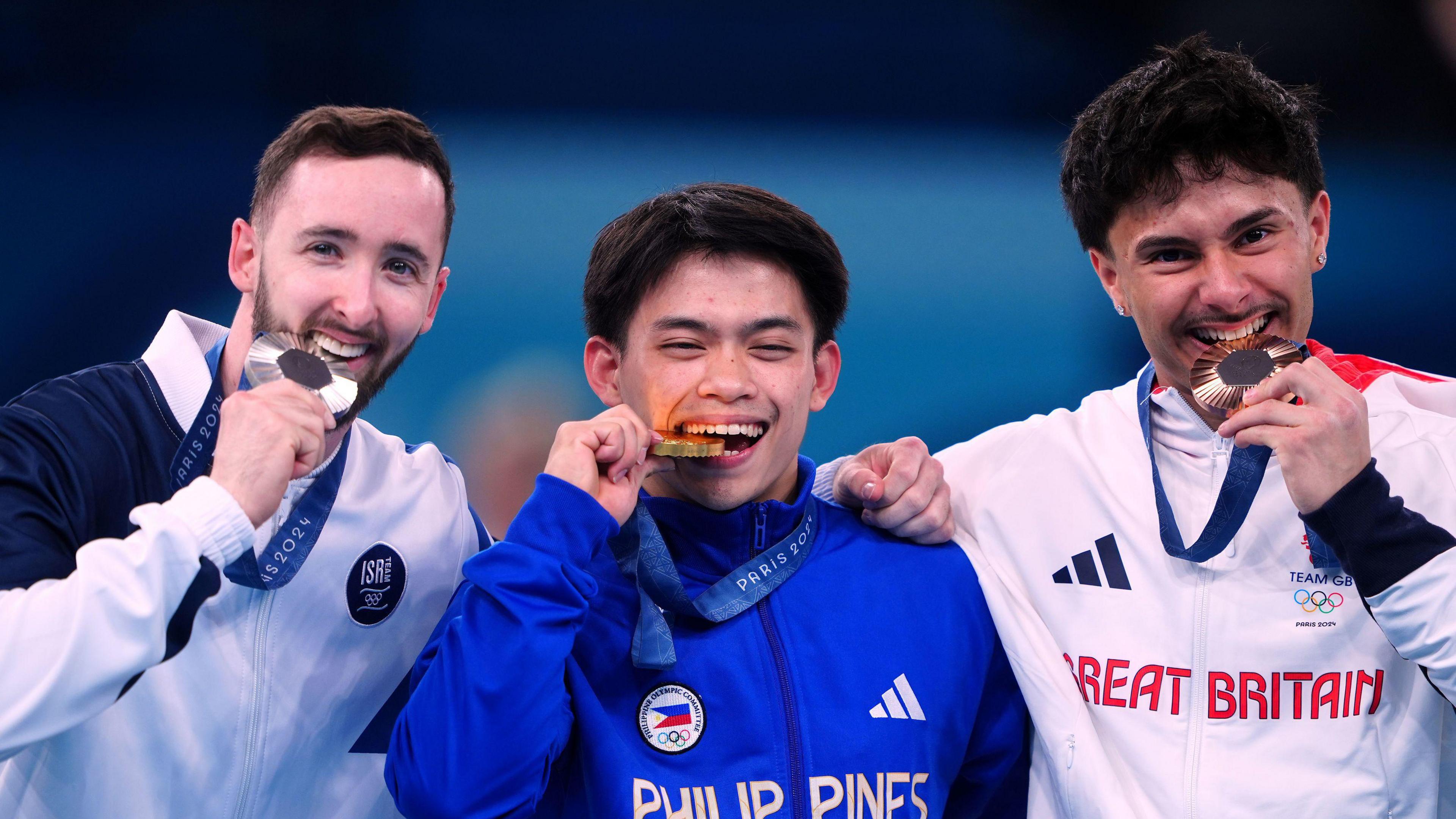 Artem Dolgopyat, dressed in the Olympic uniform of Israel, Carlos Edriel Yulo, dressed in the Olympic uniform of the Philippines and Jake Jarman in Team GB uniform, stand in a line, biting their medals and smiling at the camera