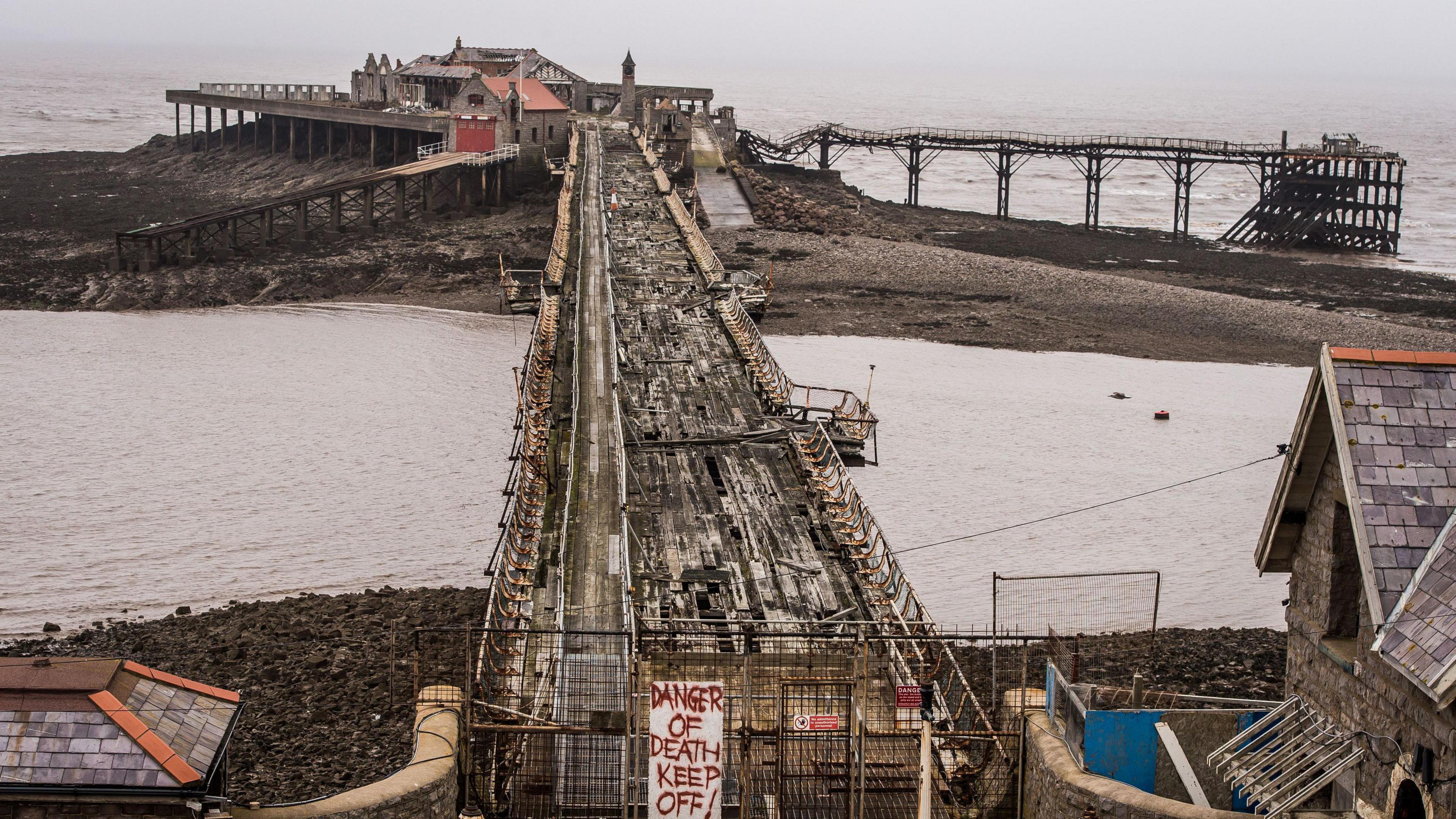 A photo taken from the shoreline of Birnbeck Pier. A sign reads 'DANGER OF DEATH KEEP OFF' in red lettering. The gangway is severely damaged, with holes in the floor and gaps in the railings. 