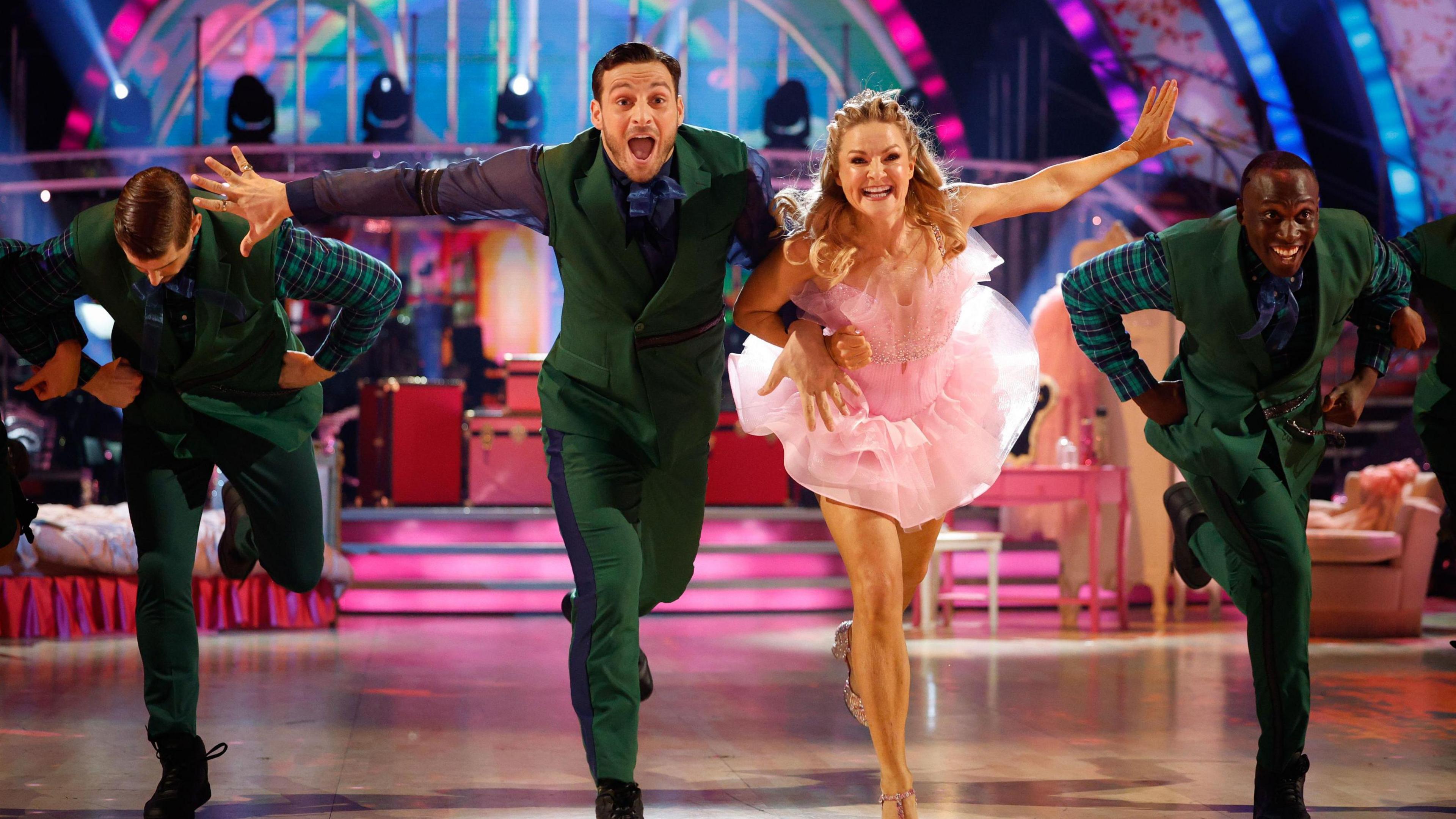  Vito Coppola in green trousers and a waistcoat smiling and throwing an arm to the side, while Sarah Hadland does the same while wearing a pink dress and interlocking her other arm with Coppola. A colourful set and a couple of other dancers are seen in the background.