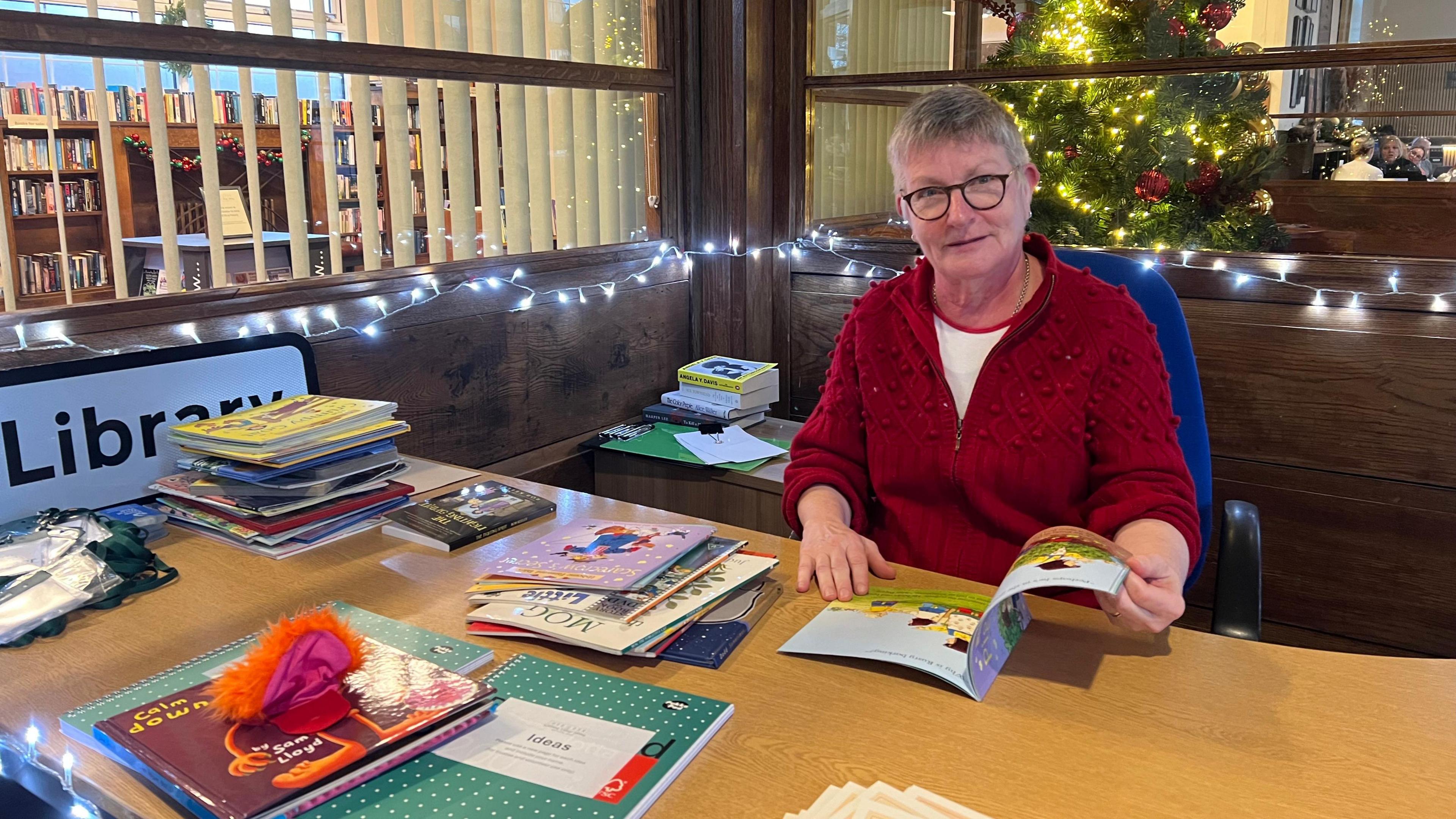 An older woman wearing a red fleece opens a book and sits at a table