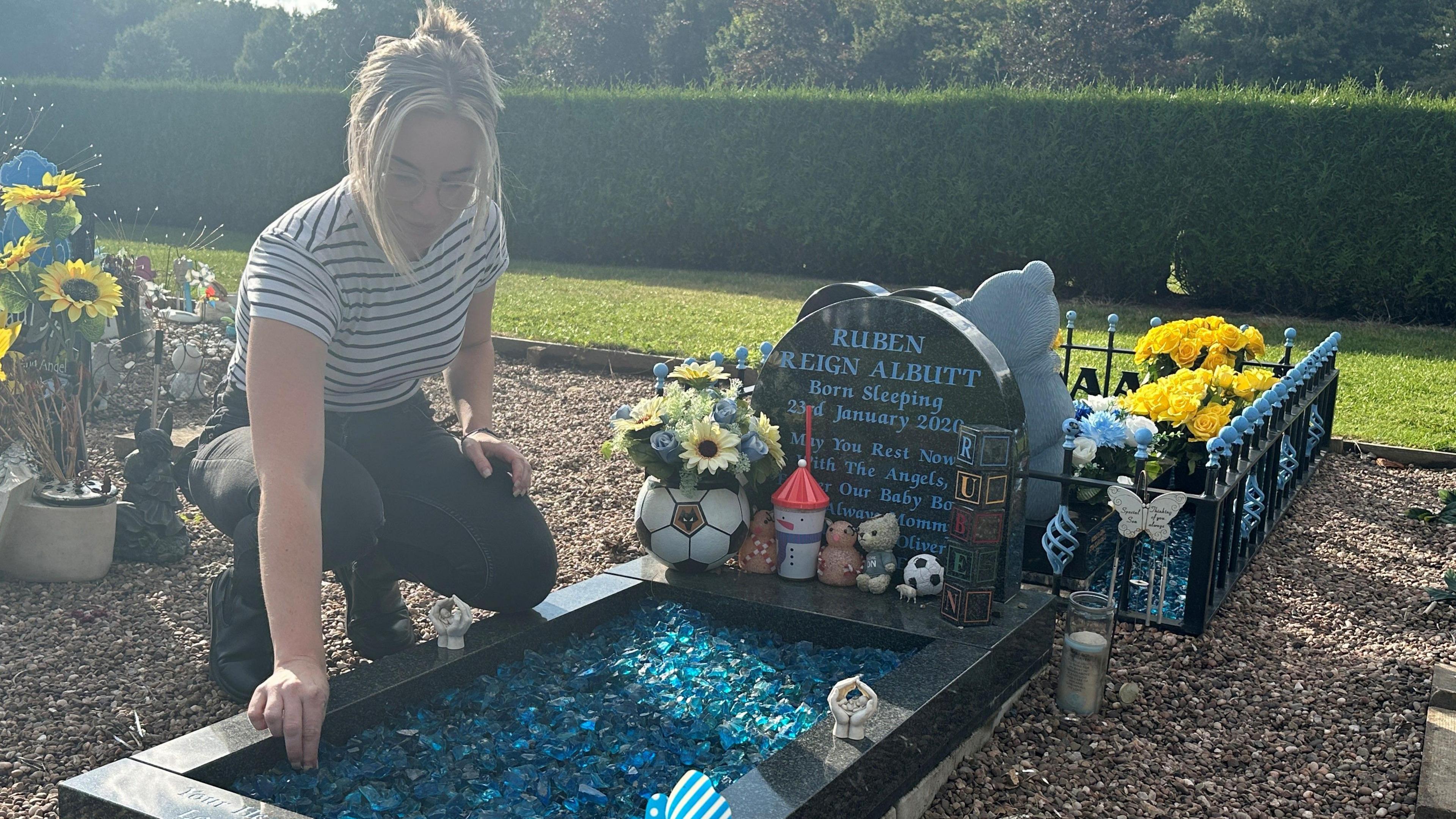 A woman in a blue and white striped t-shirt crouches above a grave. It has a headstone that says Ruben Reign Albutt, which is surrounded by toys.