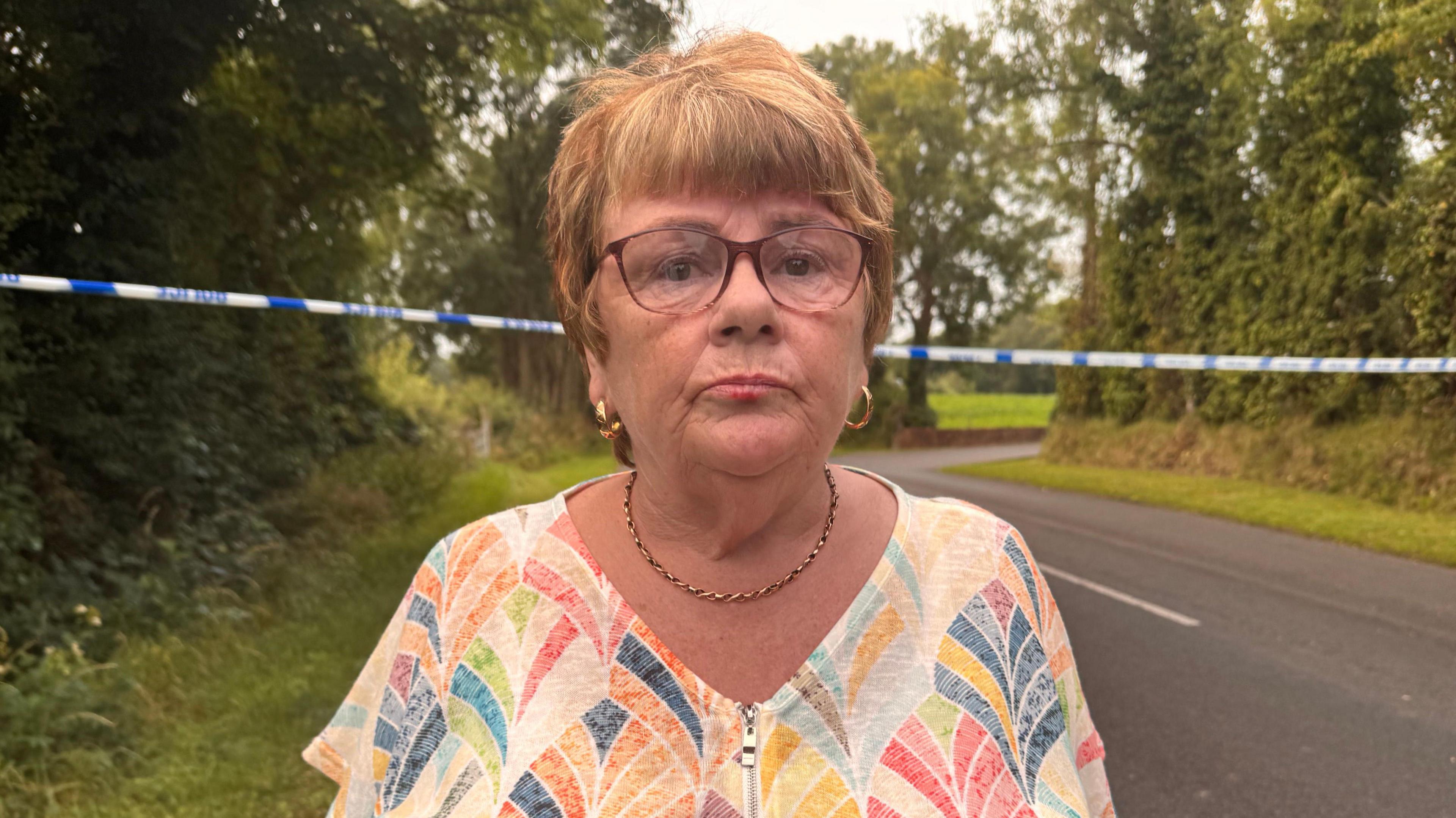 Josephine Faulkner, a woman with glasses and short, blonde hair, stands in front of a police cordon on a rural roadside. She is wearing a multi-coloured, short sleeved top, gold hooped earrings and a gold chain necklace.