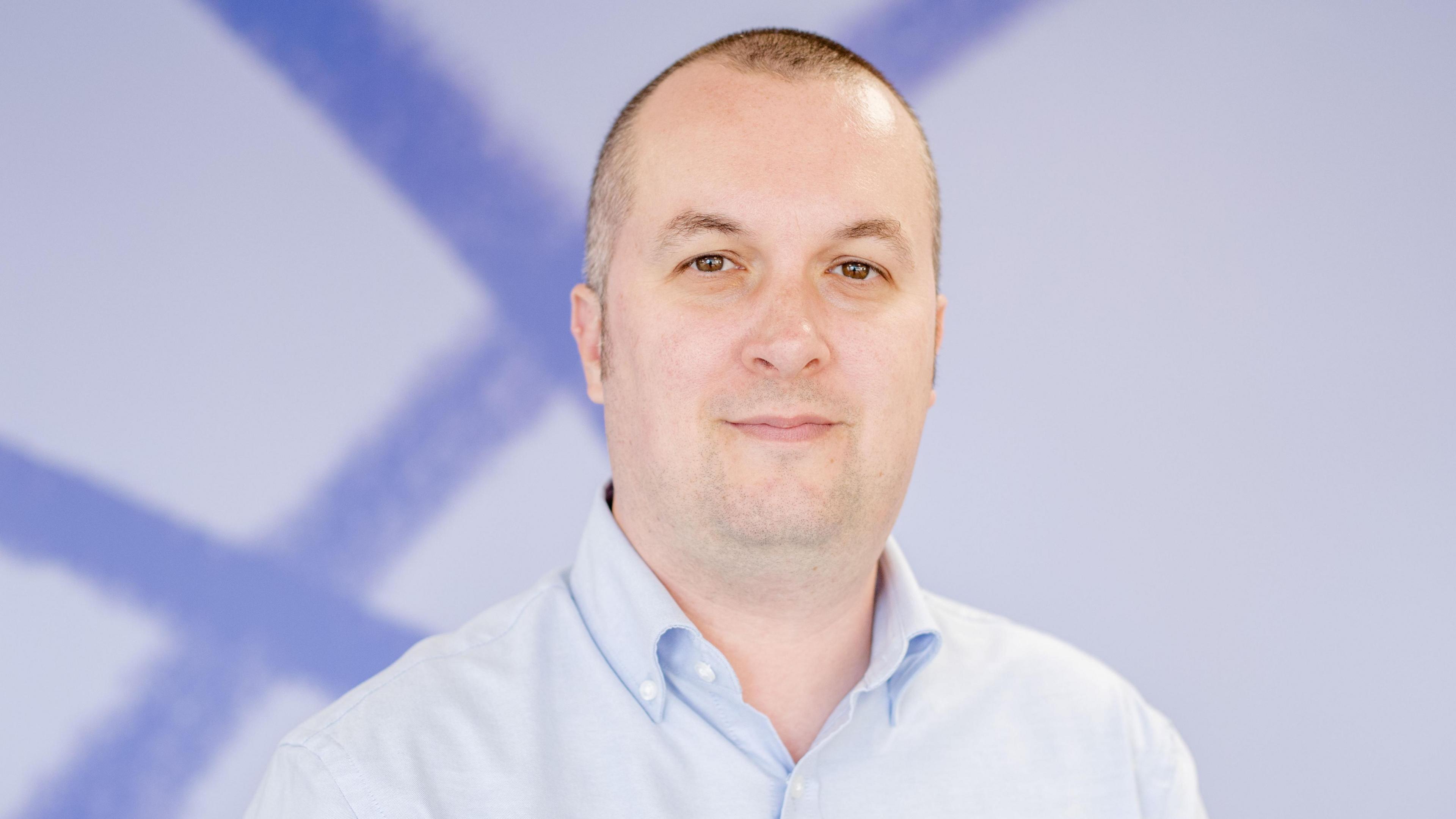 Simon Jones, a white man with shaved brown/grey hair looks into the camera, smiling slightly. He wears a light blue shirt and stands in front of a white and blue background.