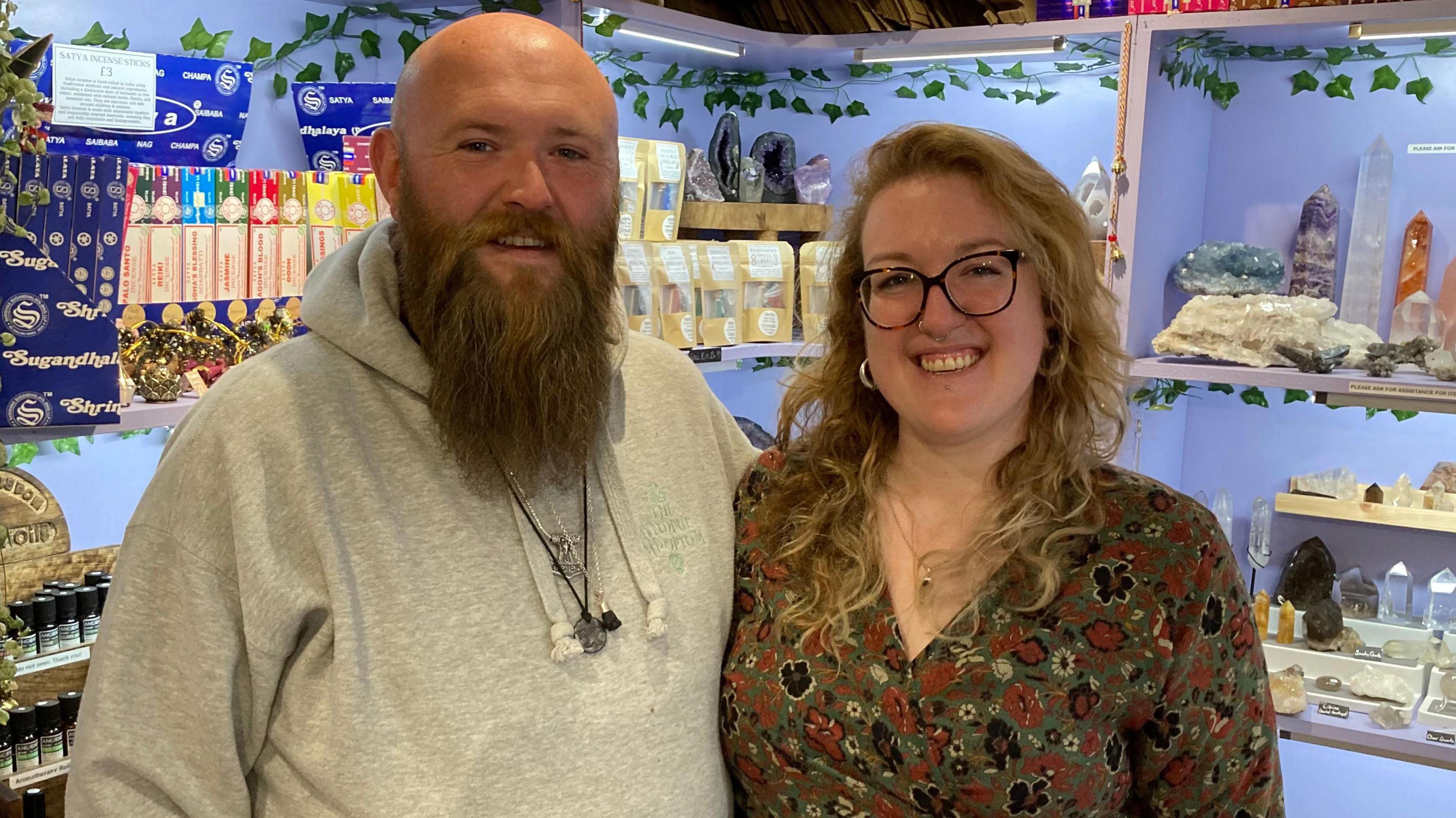 A man with a beard and bald head smiles toward the camera, he is wearing a great hooded top , standing next to him is a woman with blond hair and wearng glasses, she is smiling toward the camera and is wearing a green dress with a brown floral pattern. Behind them are crystals and minerals on display on shelving units.