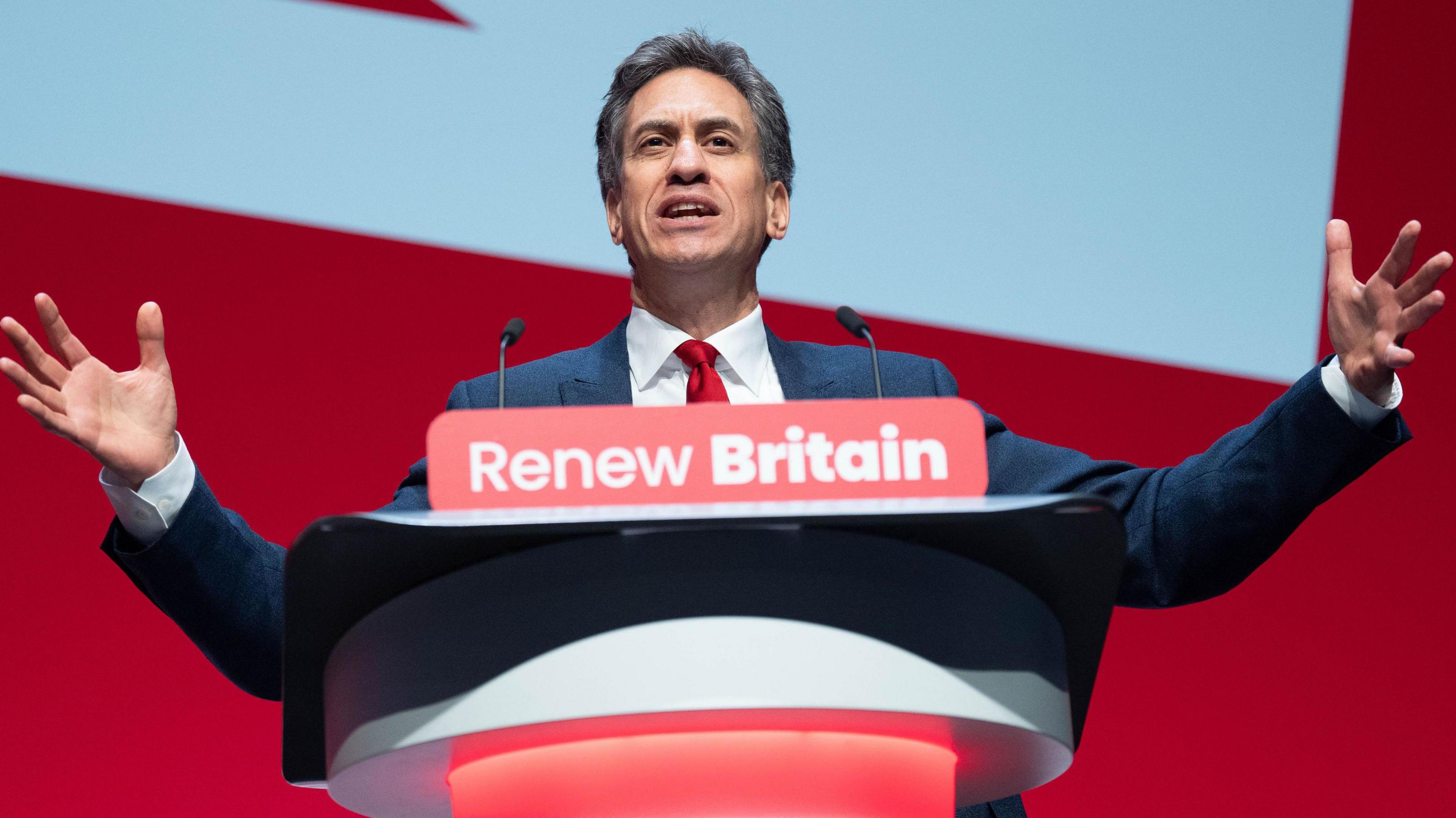 A man with dark hair is standing at a podium with the words Renew Britain written on the front of it. He has his arms outstretched. He is wearing a navy suit with a white shirt and red tie. The background is a red and blue screen.
