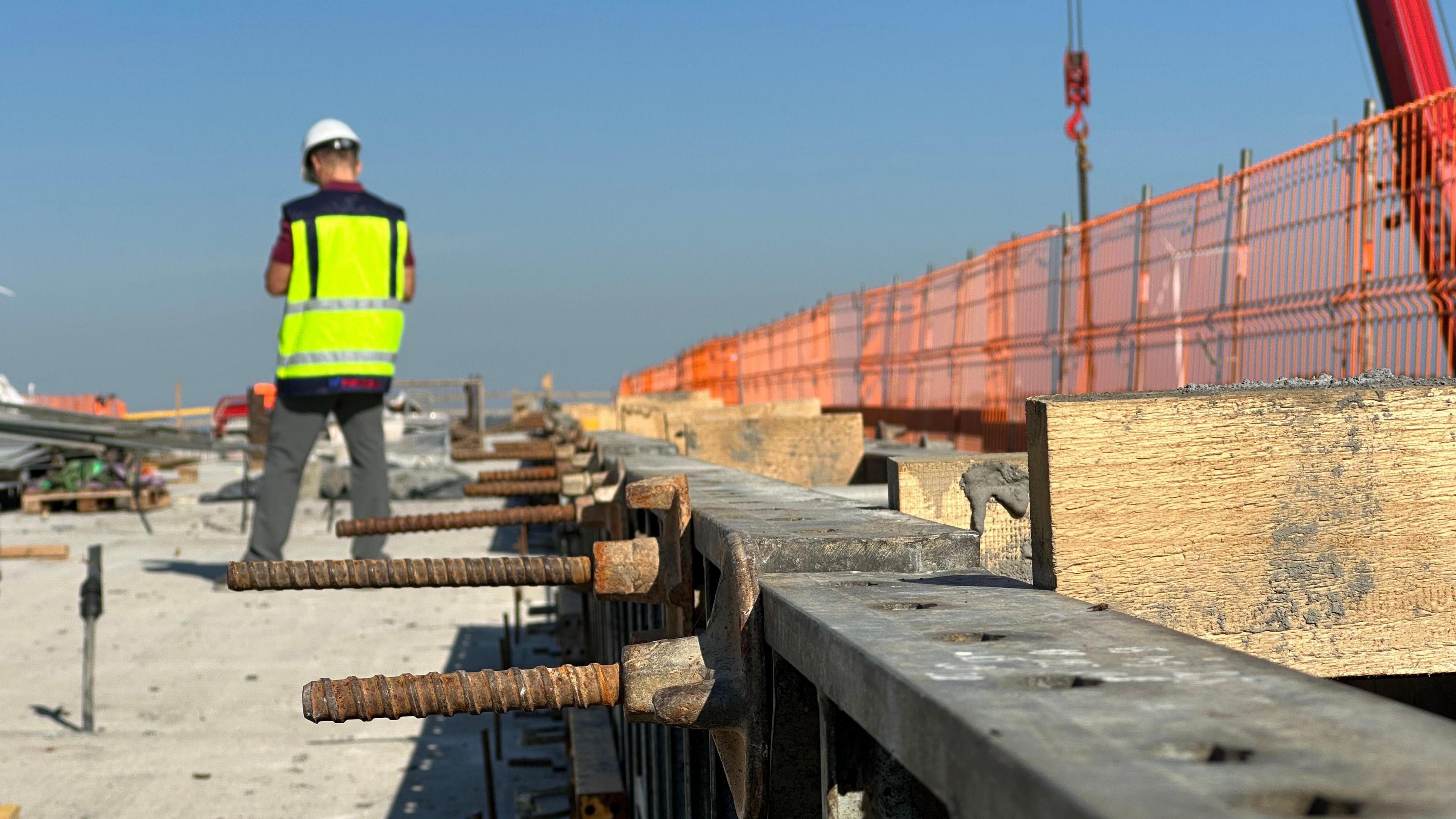 A construction worker in a neon yellow vest and white hard hat stands beside concrete with screws sticking out of it as he works on the new rail project in Lithuania