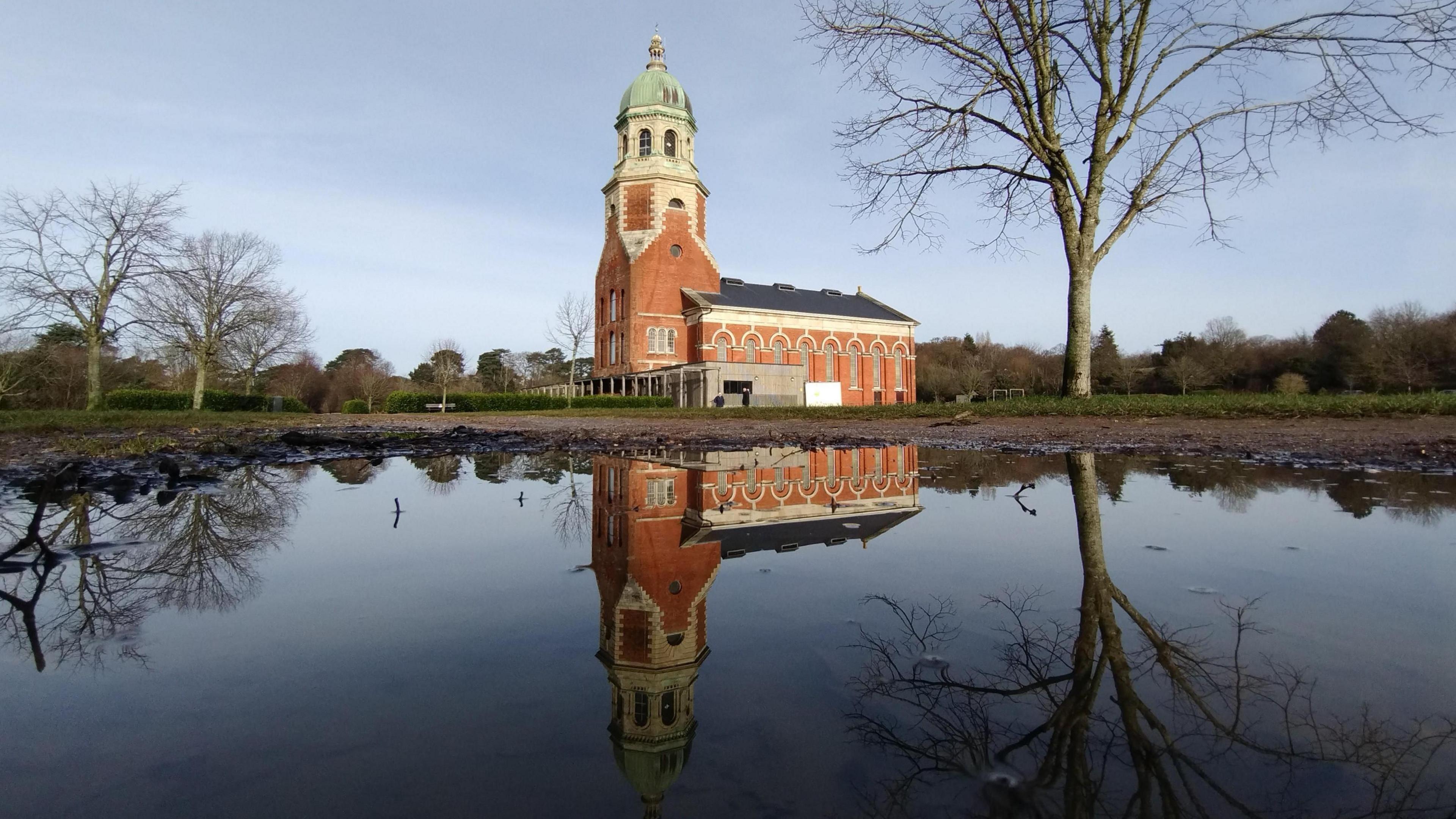 The chapel in the Royal Victoria Country Park in Netley is reflected in a puddle. The red brick building has a spire. It is surrounded by a grass area with several trees. The sky is overcast. In the foreground the scene is reflected in a puddle on a path.