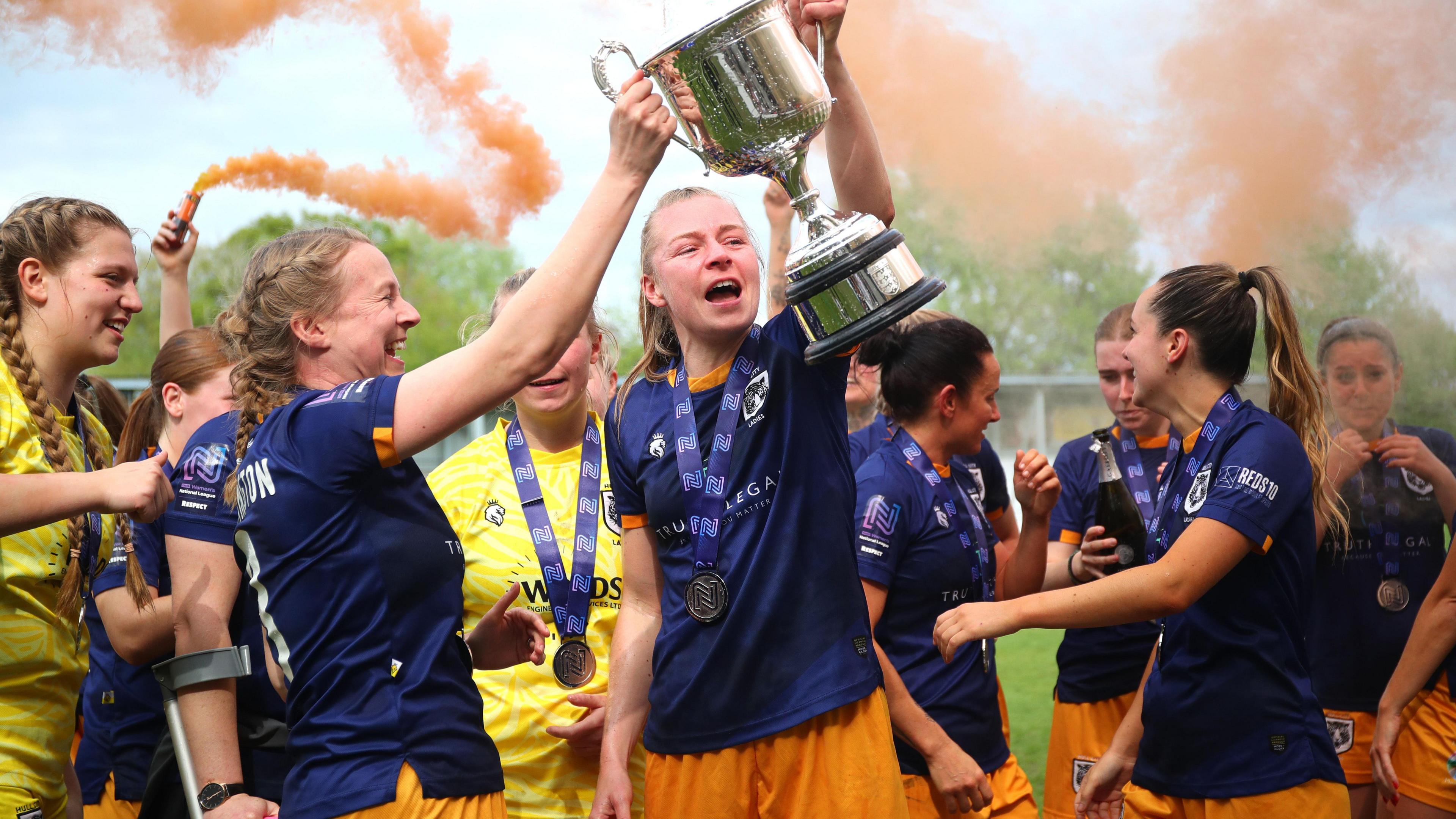 A group of 10 women, most wearing navy blue football shirts and amber shorts, with two in yellow tops, smile as they celebrate winning a league title. Two hold up a silver trophy between them. pose for a selfie with a silver trophy as amber-coloured flares go off behind them. They have medals around their necks.