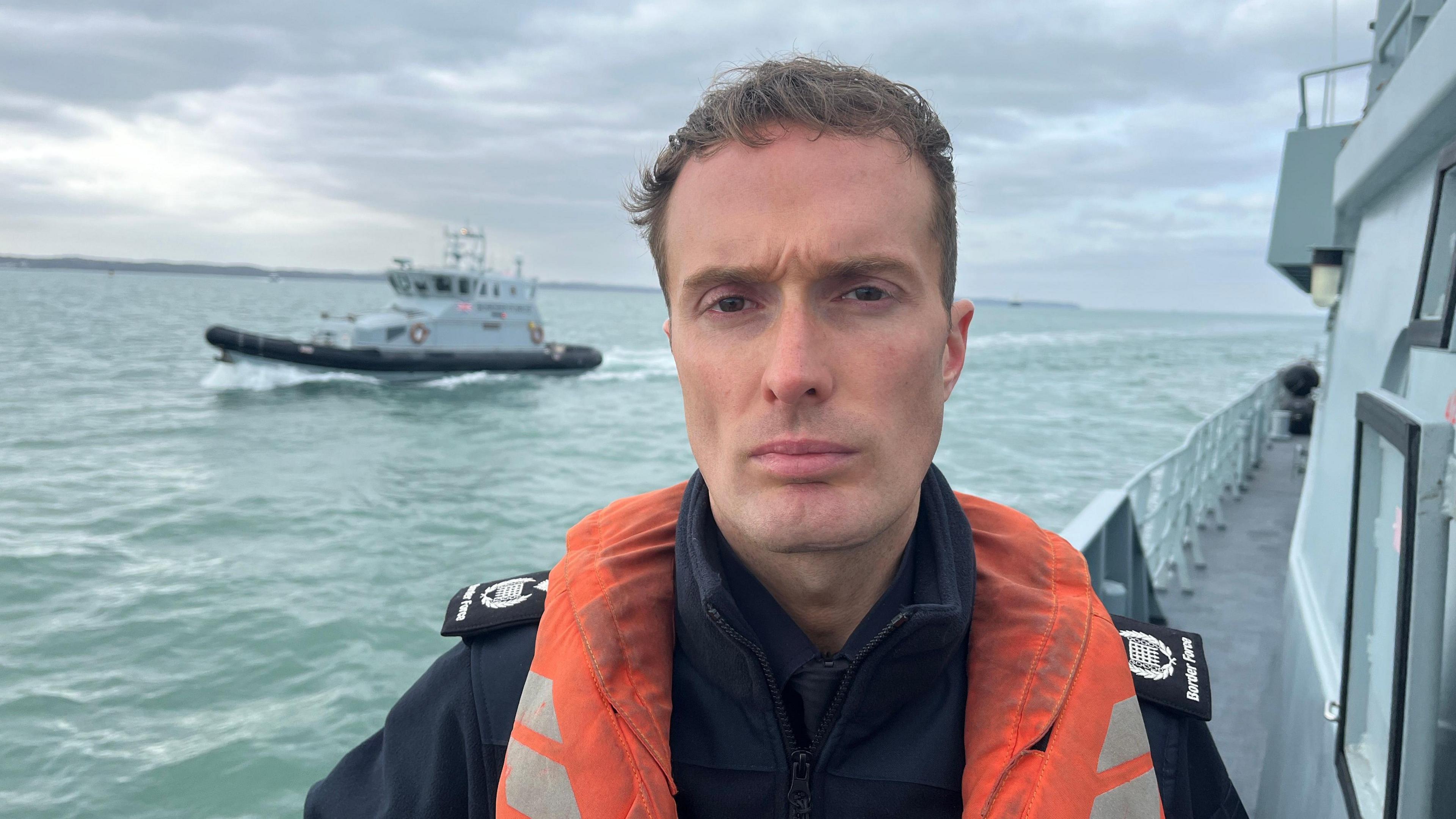 A uniformed officer, standing on the deck of a Border Force cutter, wearing a lifejacket and staring solemnly into camera. A second Border Force vessel is at sea in the background.