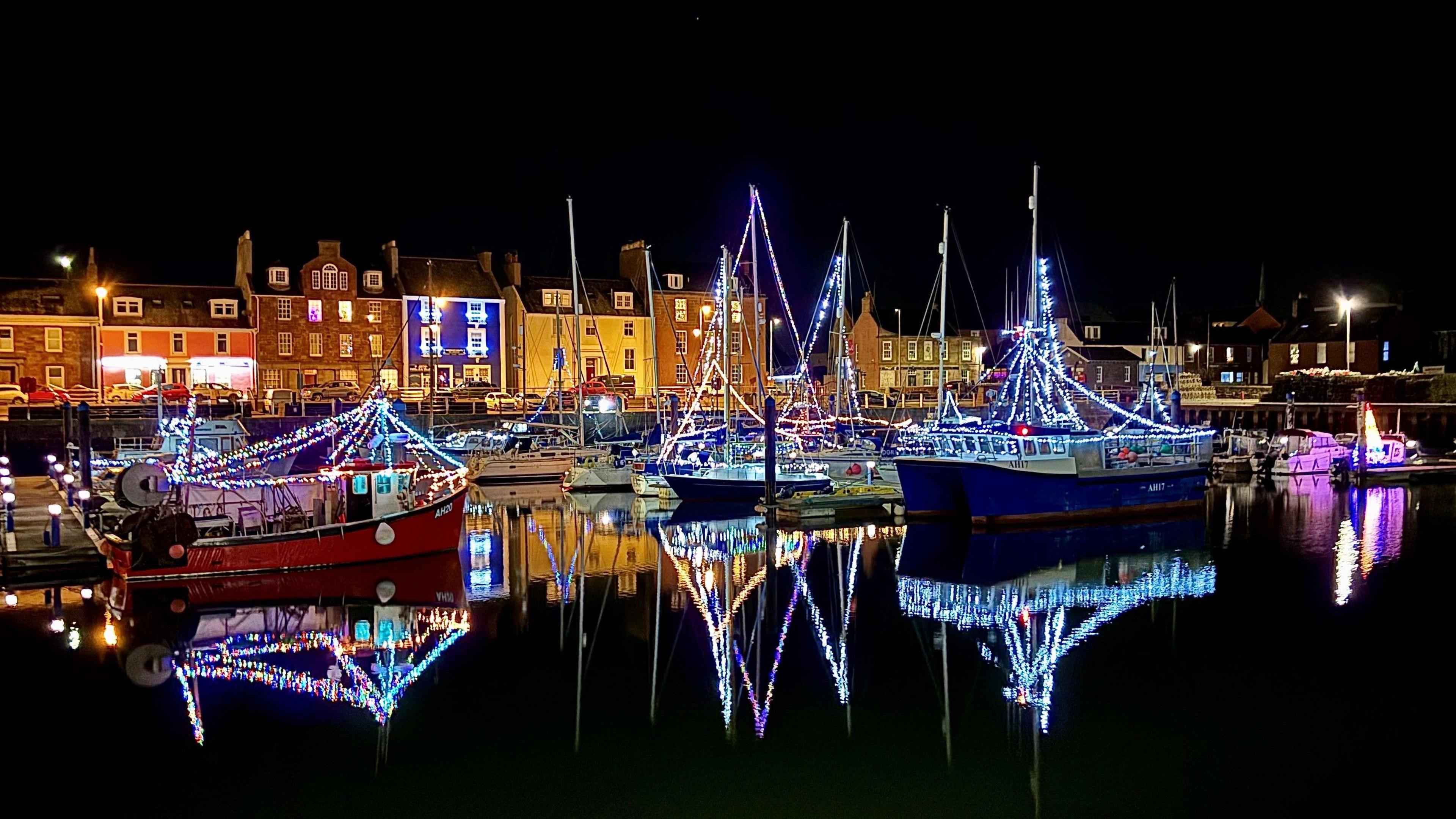 A number of boats in the harbour at Arbroath. It is dark. The boats have multi-coloured Christmas Lights around their masts and rigging. The lights are reflected in the dark water below. In the background is a row of multi-coloured houses. Most of them have their lights on. The sky overhead is also dark.