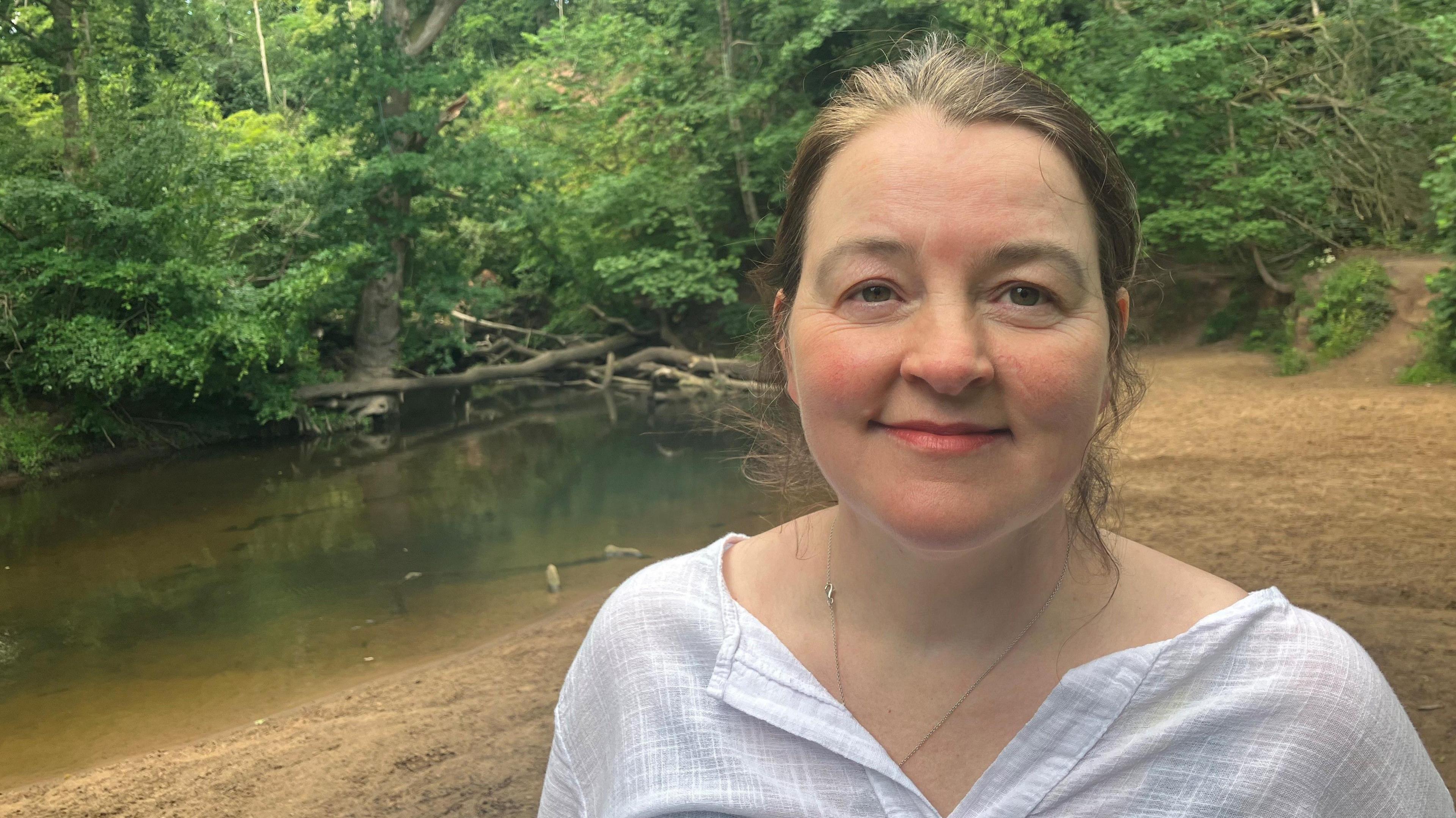 Photograph of Lisa Smith from Handforth. She has light brown hair tied back and is smiling for the camera while standing on the banks of the River Bollin on the Quarry Bank Mill estate.