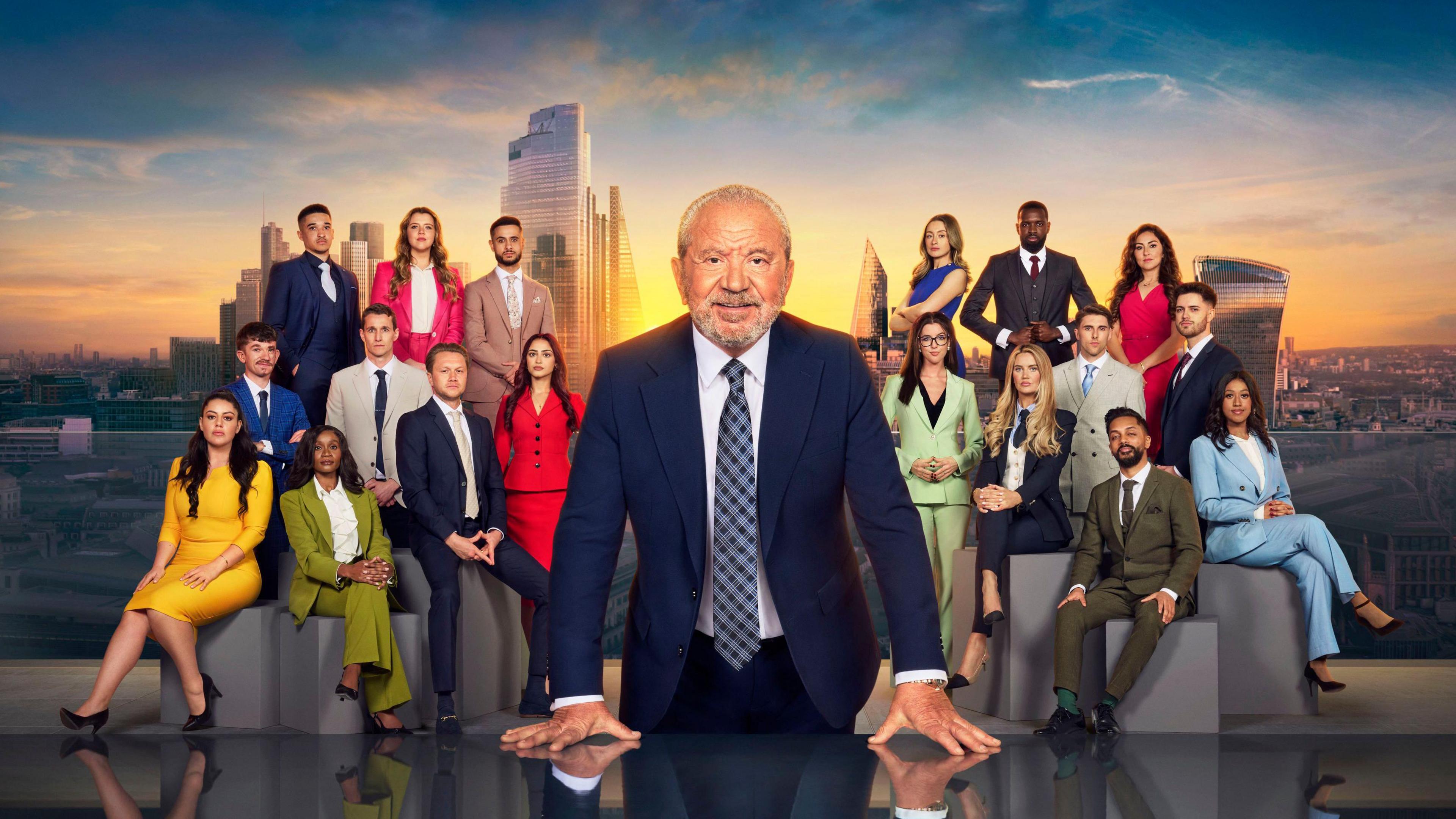 Lord Sugar stands in front of the 18 candidates for series 19 of the Apprentice, with a sunlit London skyline behind all of them.