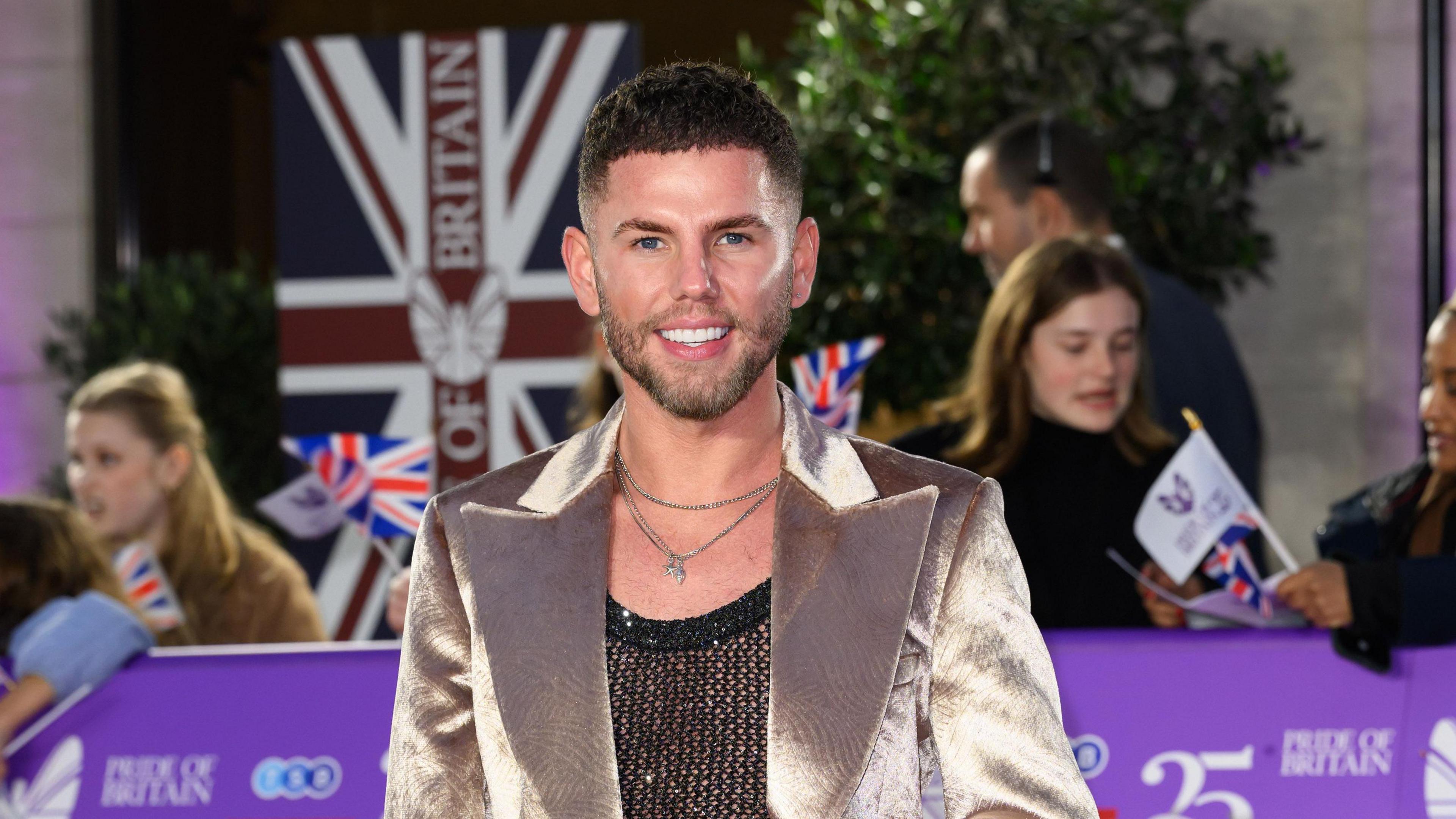 Dean McCullough wearing a shiny blazer and smiling at camera. A Union Jake can be seen flying in the background