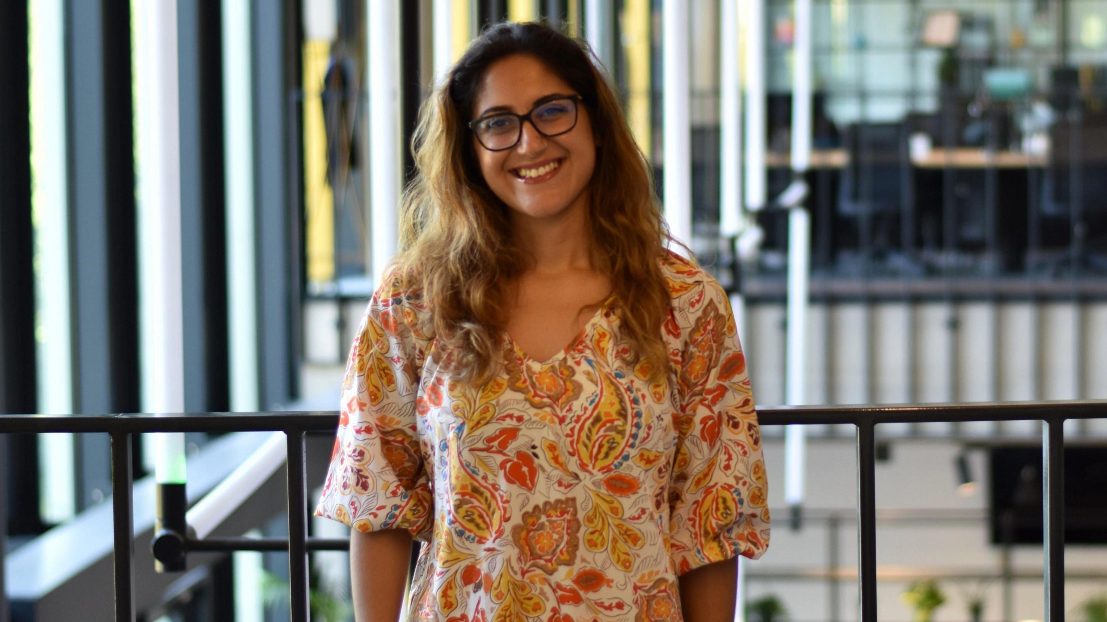 Dr Layla Hosseini-Gerami is smiling at the camera in the atrium of a large office building. She is wearing a multicoloured patterned dress and glasses.