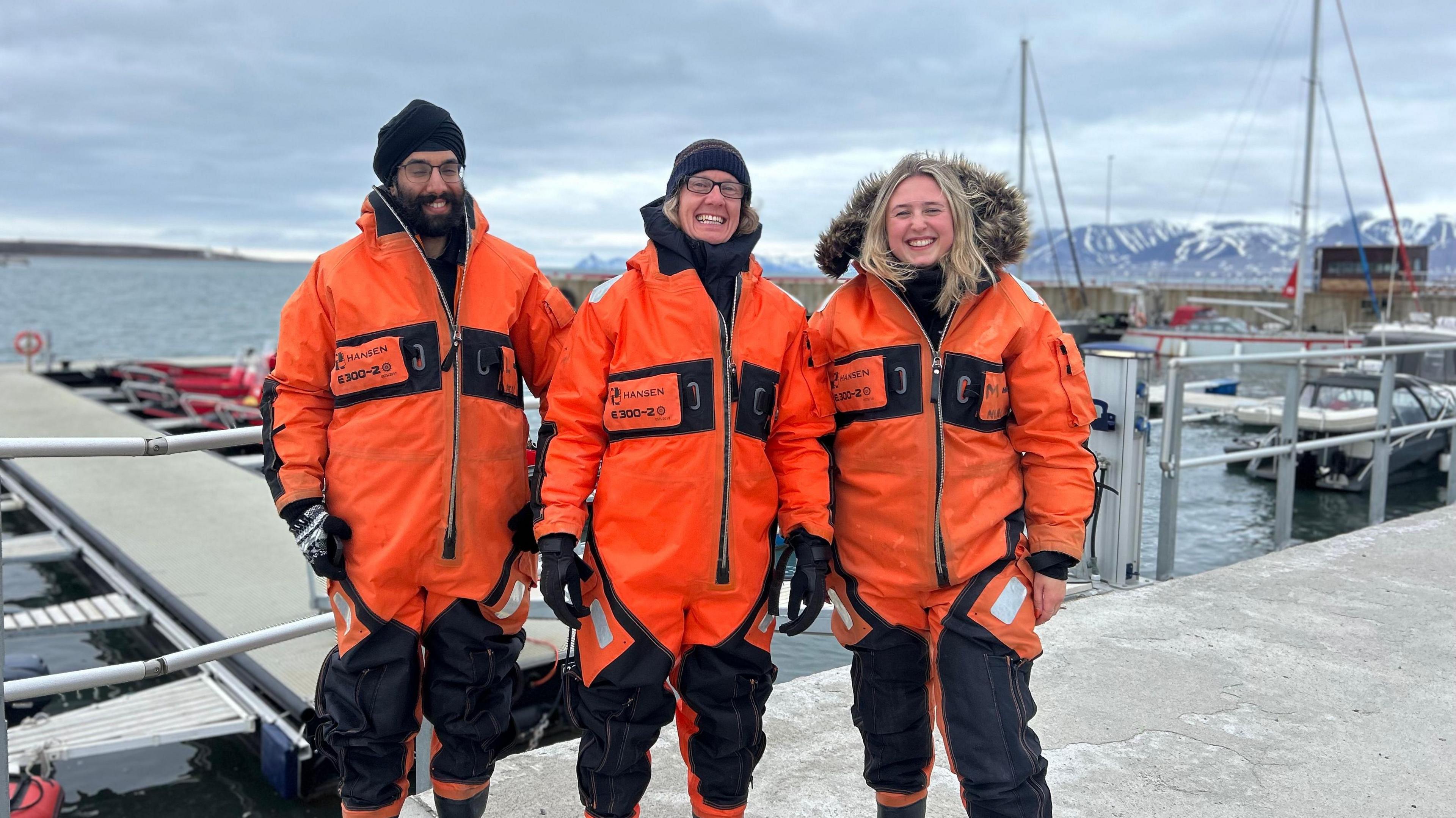 Three researchers from the University of Leicester on their arctic voyage wearing orange overalls. Stood in front of a concrete jetty. Snow-capped mountains are in the background.