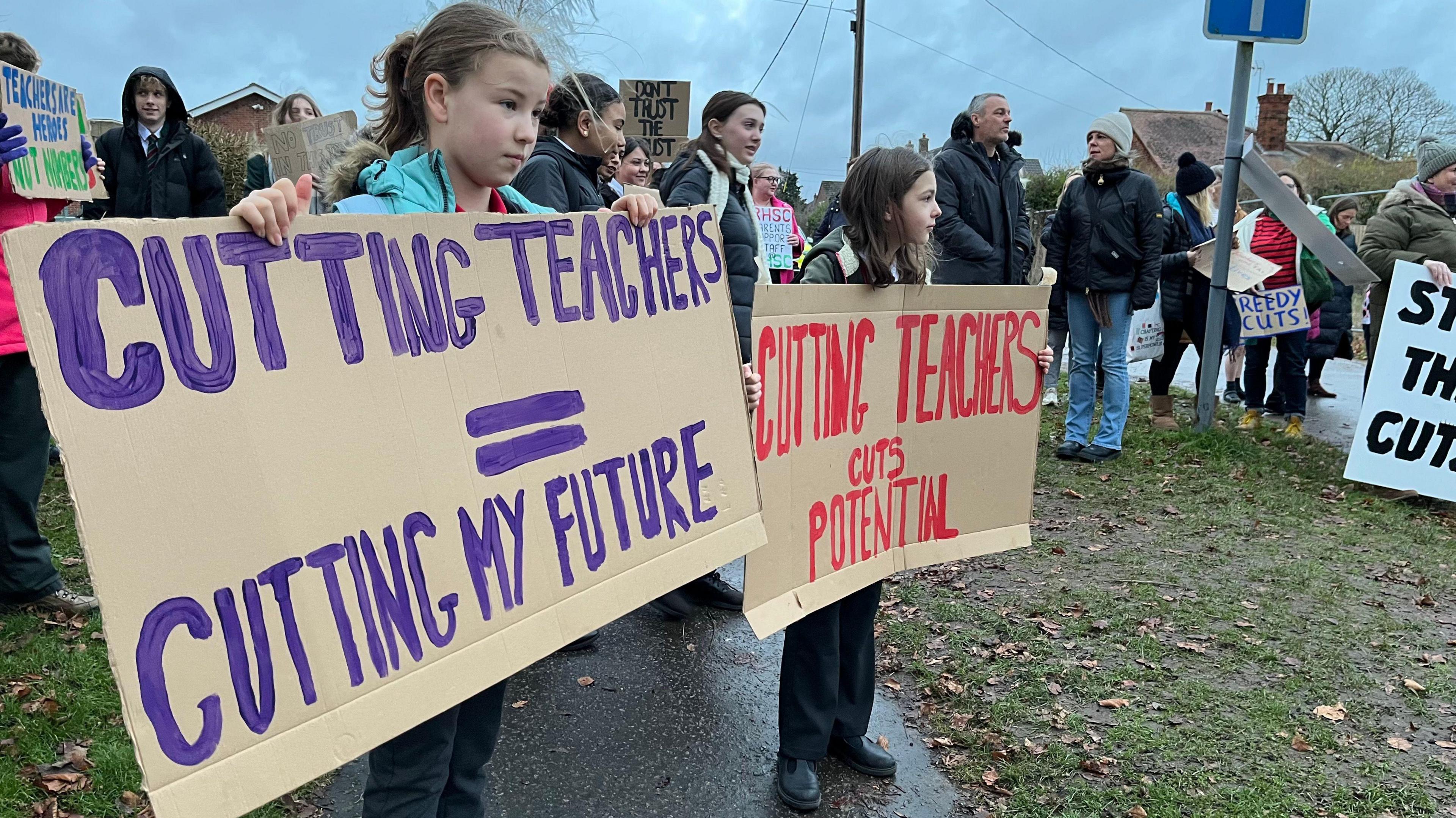 Adults and children gather on a street. It is cold and they are wearing warm clothing. Two children in the foreground are holding cardboard placards.