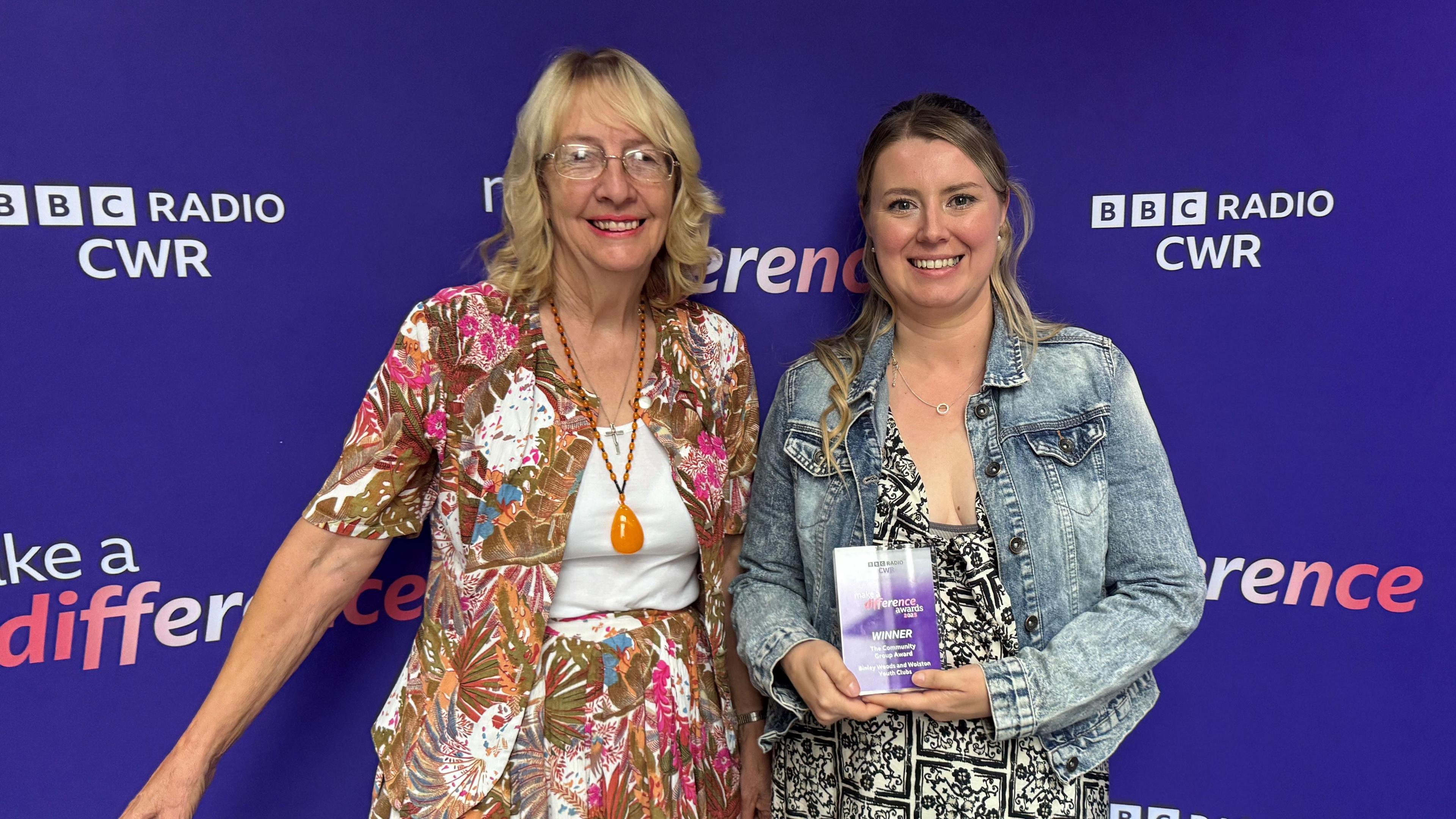 Two women - Emma Stuchbury and Shan Dobinson - are standing in front of a purple BBC CWR backdrop, holding a purple trophy.
