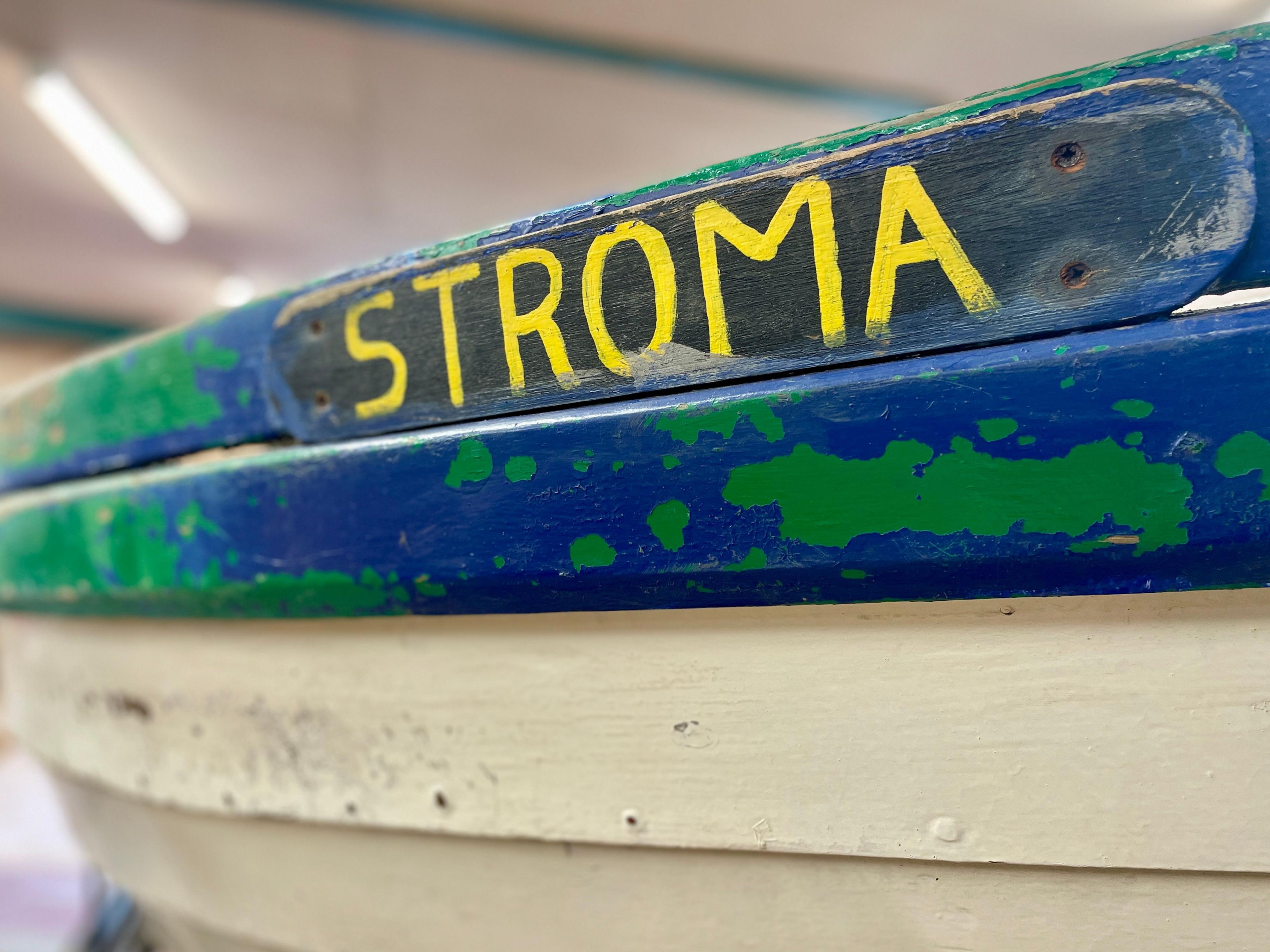 A black nameplate with yellow writing 'Stroma' attached to the hull of a blue and white boat with flaking green paint in places