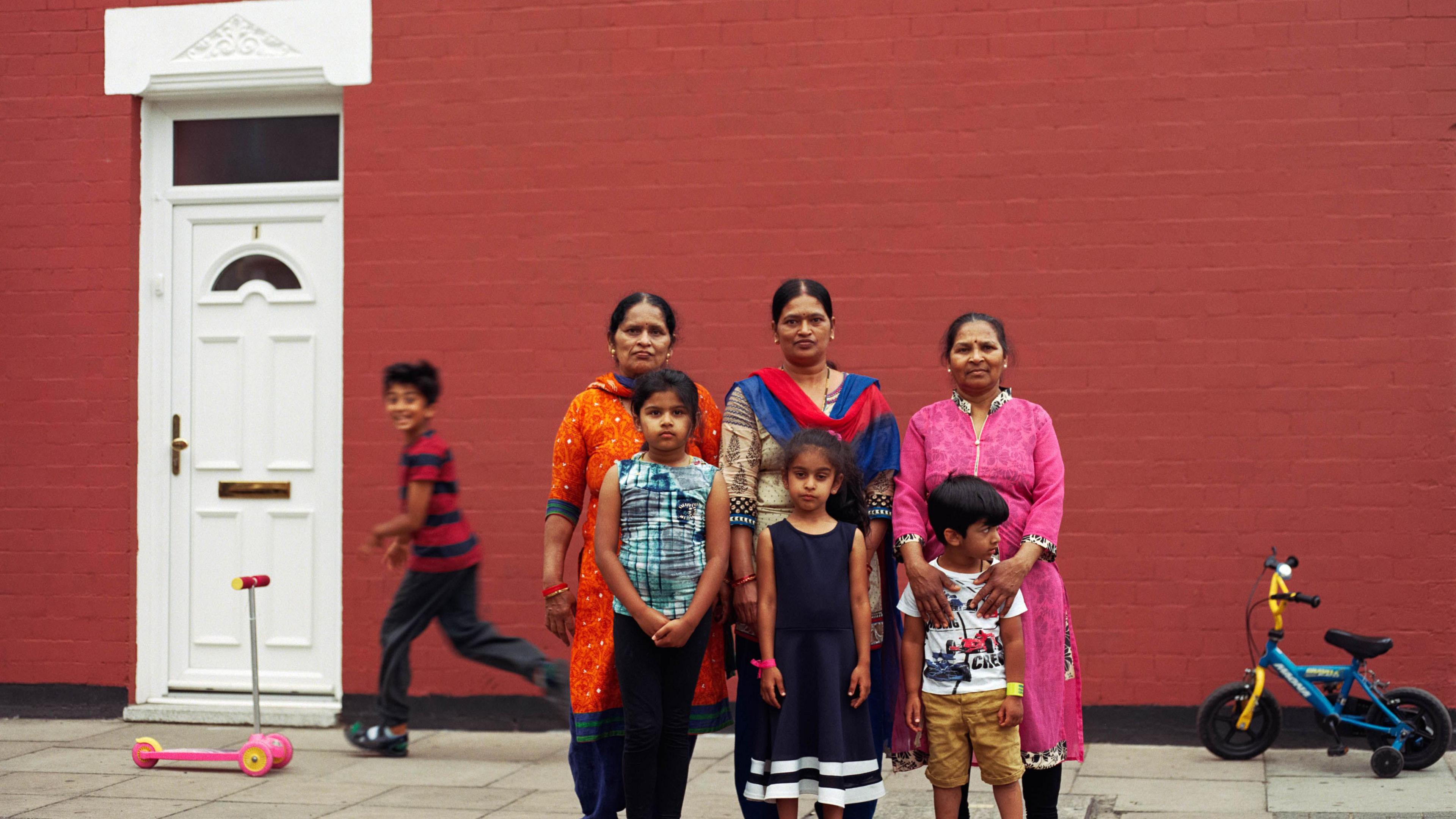 Three women pose in front of a house with three children arranged in front of them and a fourth child running past with a smile on his face