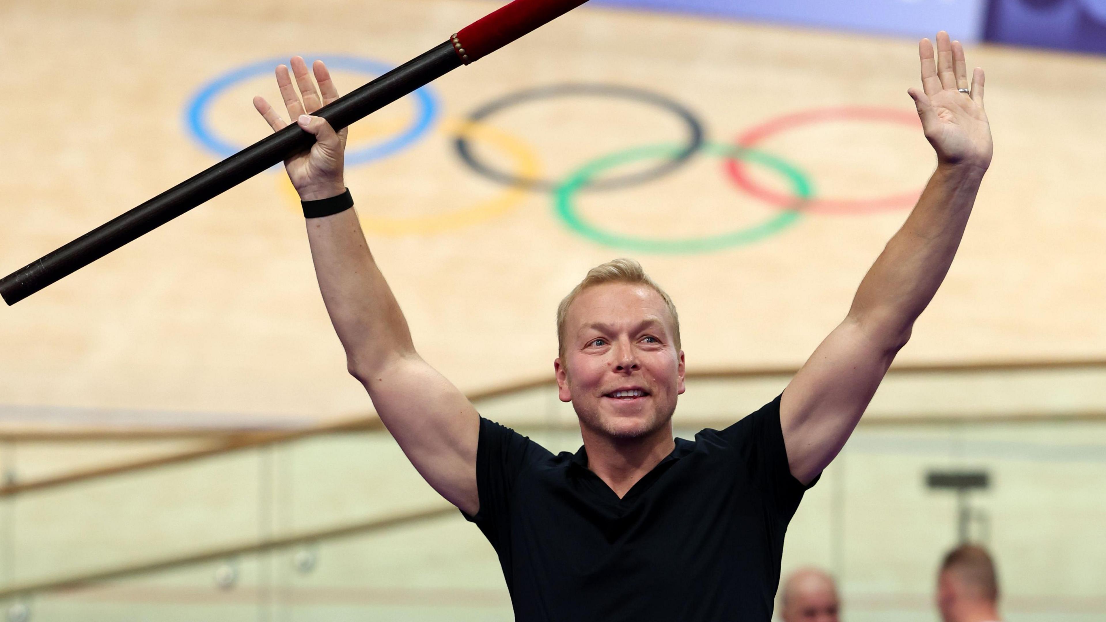 Sir Chris Hoy, a man with short hair and a black t-shirt, stands in front of a wooden velodrome track with the Olympic rings on it. He is smiling and is holding up his hands, one of which is holding a baton