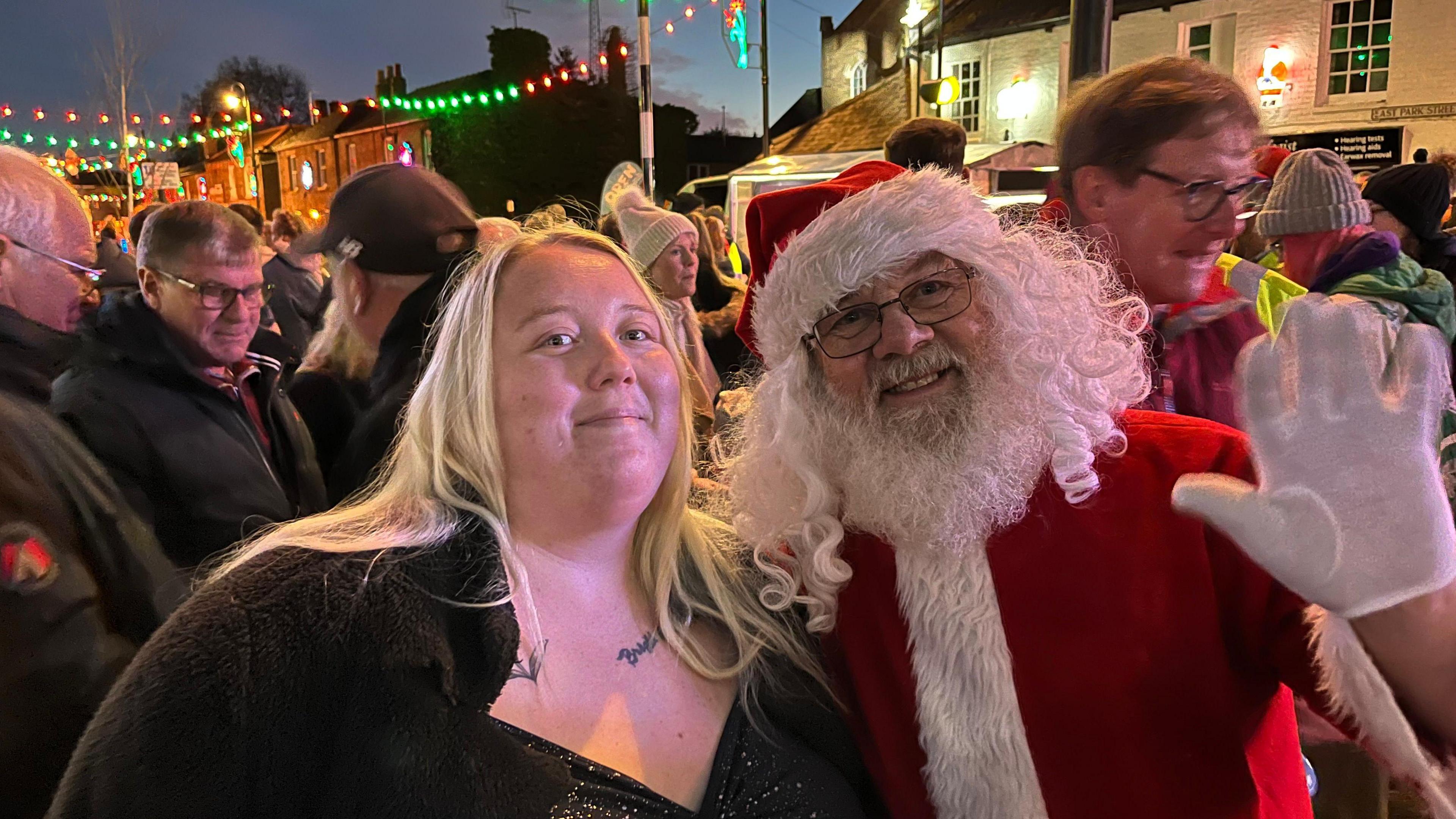 Devon Mandley, 32, has long blonde hair which is parted. She is wearing a black sparkly dress with a black cardigan on top. Some tattoos are visible near her neck. She is standing next to someone dressed as Santa, who is waving. Behind is a large crowd of people in the street with colourful lights strung over the road.