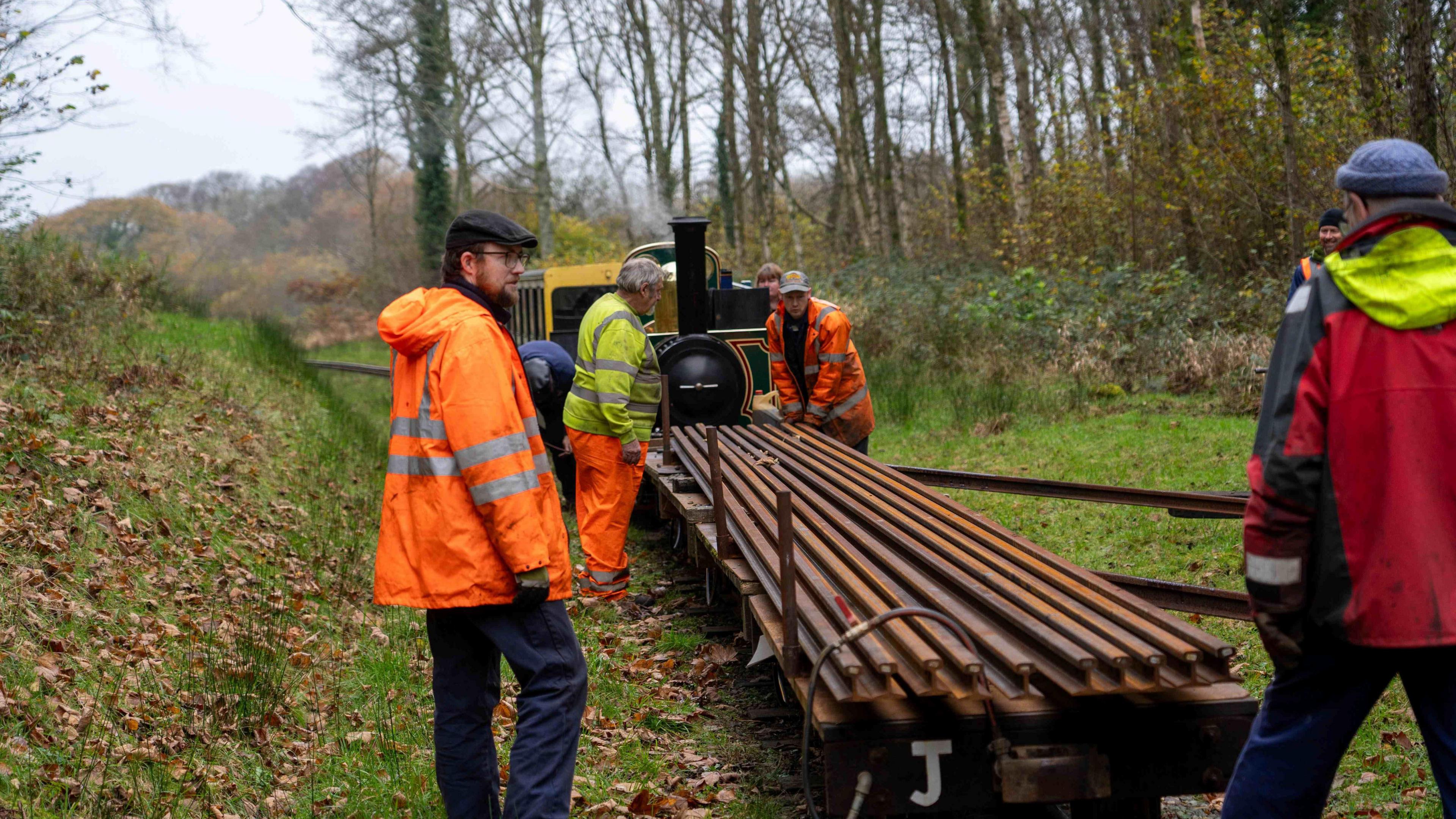 Workmen load materials onto a flat trailer attached to a steam train. The train is pictured from behind with workmen around it. They are within woods along the track.