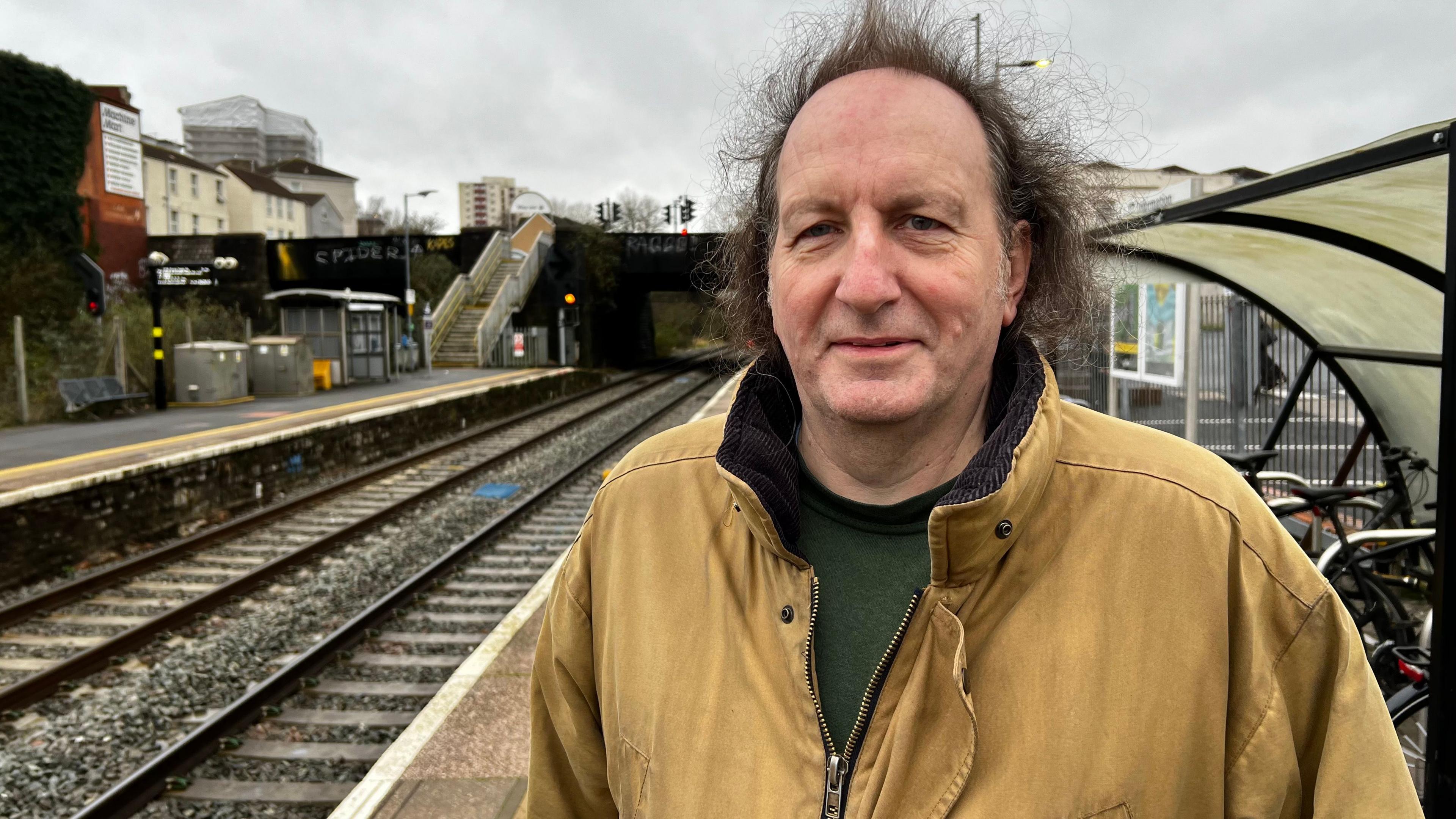 Bruce Williamson stands at a train station platform on a gloomy overcast day. He has long, dark brown hair and wears a dark green jumper underneath a thick brown jacket.