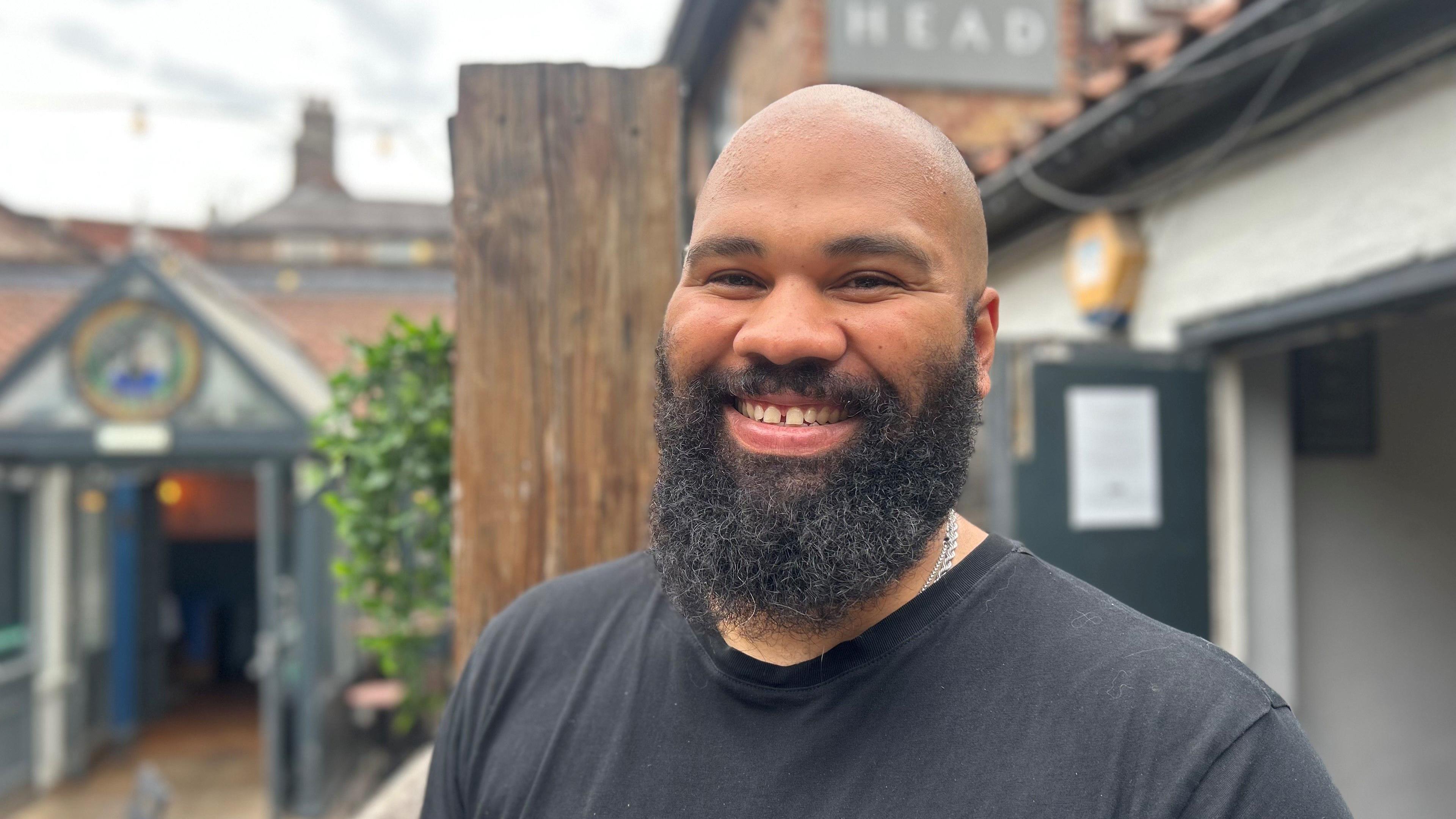 Aiden Hazlewood, who has a bald head and black beards, smiles. Behind him is The King's Head pub